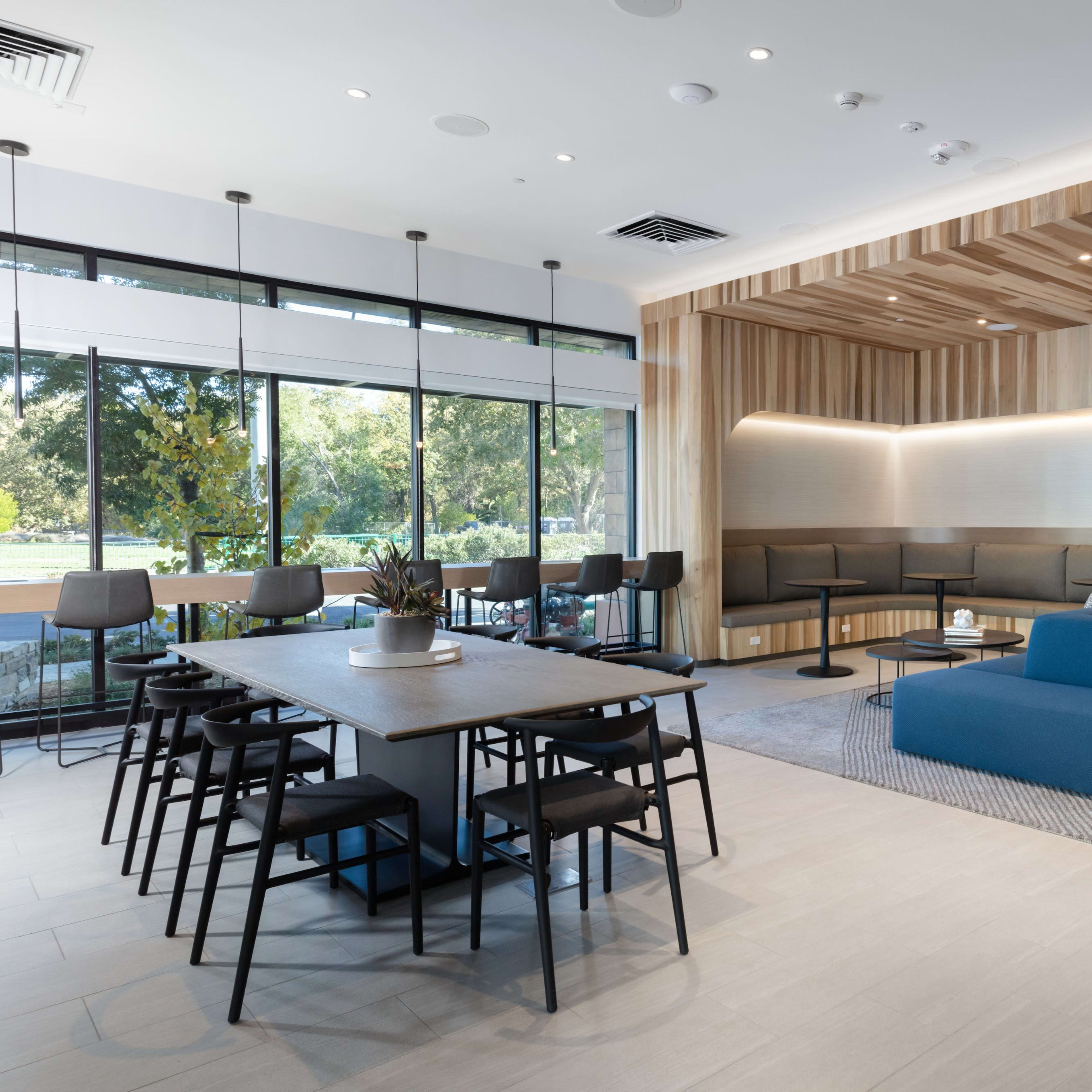 A modern lounge area features a blue sofa, a wooden accent wall, tables with chairs, and large windows allowing natural light.