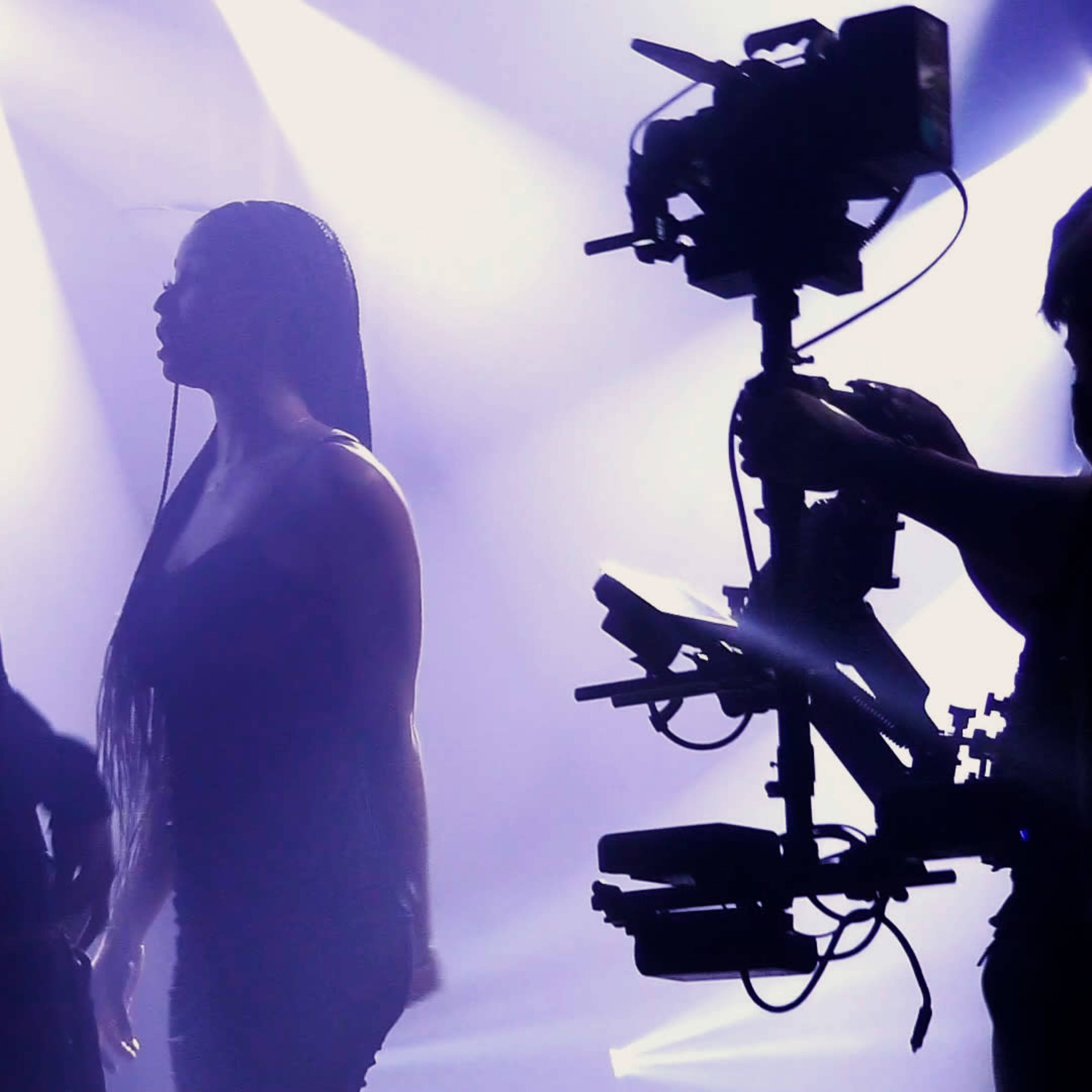A man and a woman stand facing each other under bright stage lights, with a camera filming them from the side.