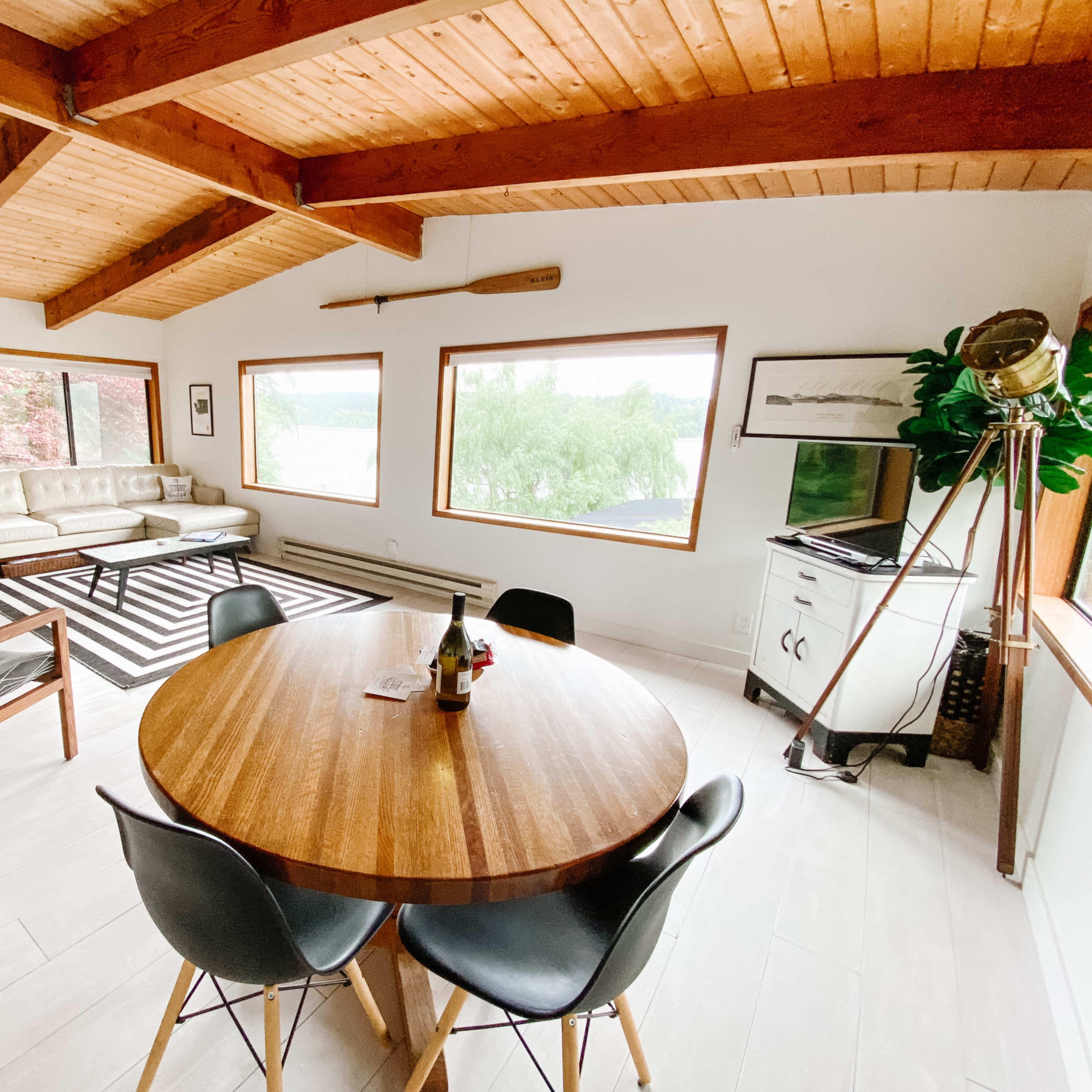 A modern living space featuring a round wooden dining table, black chairs, a contemporary sofa, large windows with views of greenery, and a wood stove.