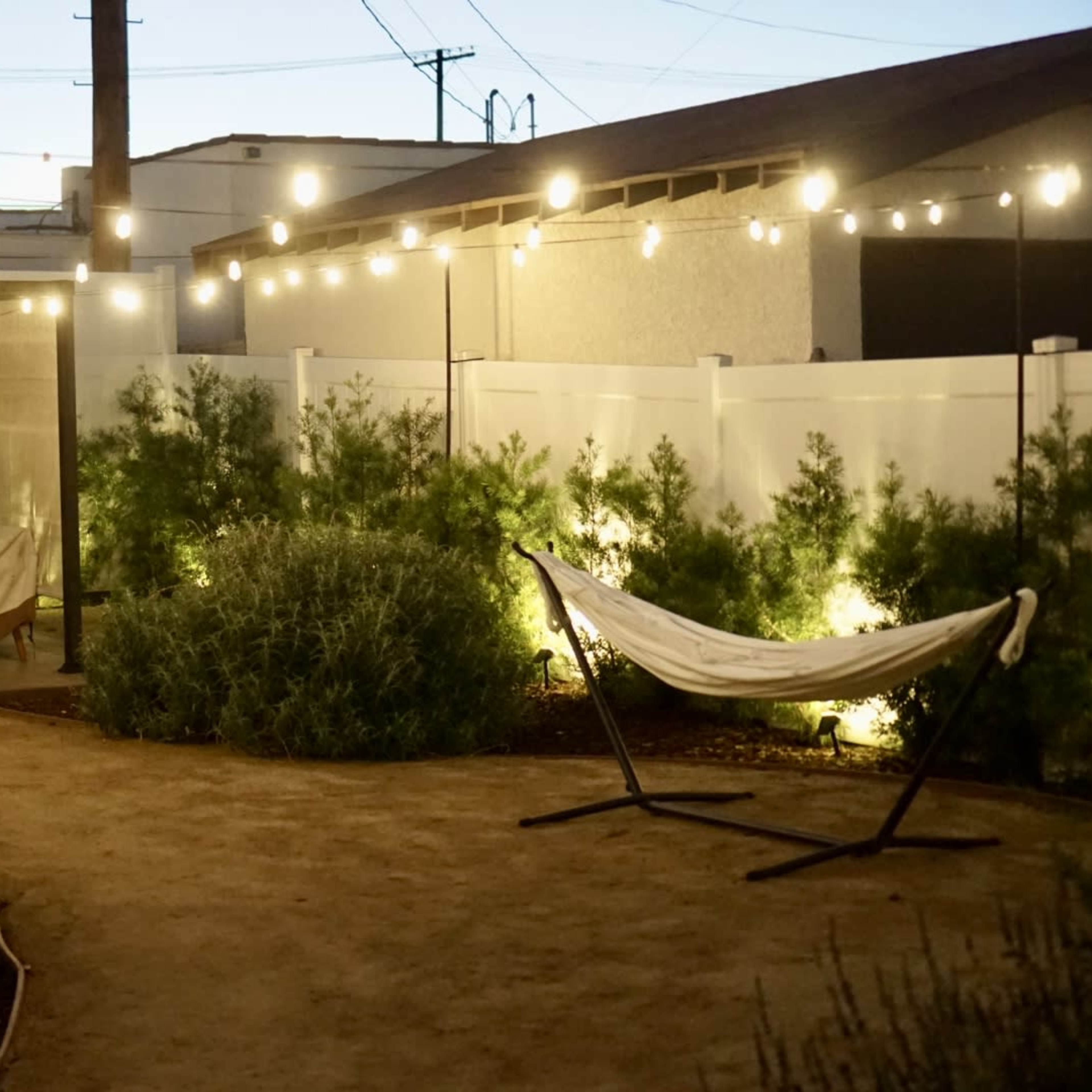 A well-lit outdoor space features a hammock, surrounded by low bushes and string lights overhead.