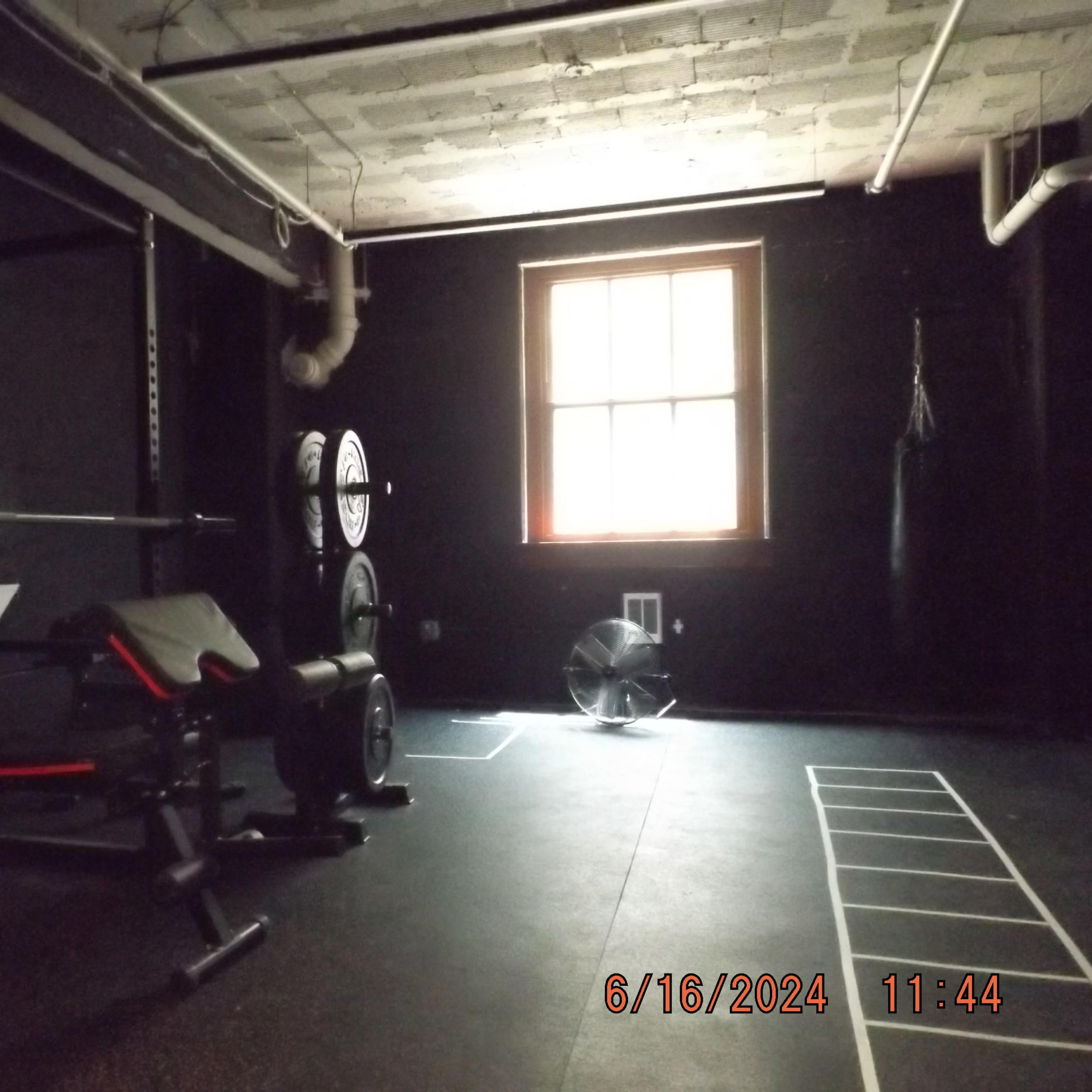 A dimly lit gym with a squat rack, weights, a bench, and a fan, alongside a wall marked with a large letter "Z."