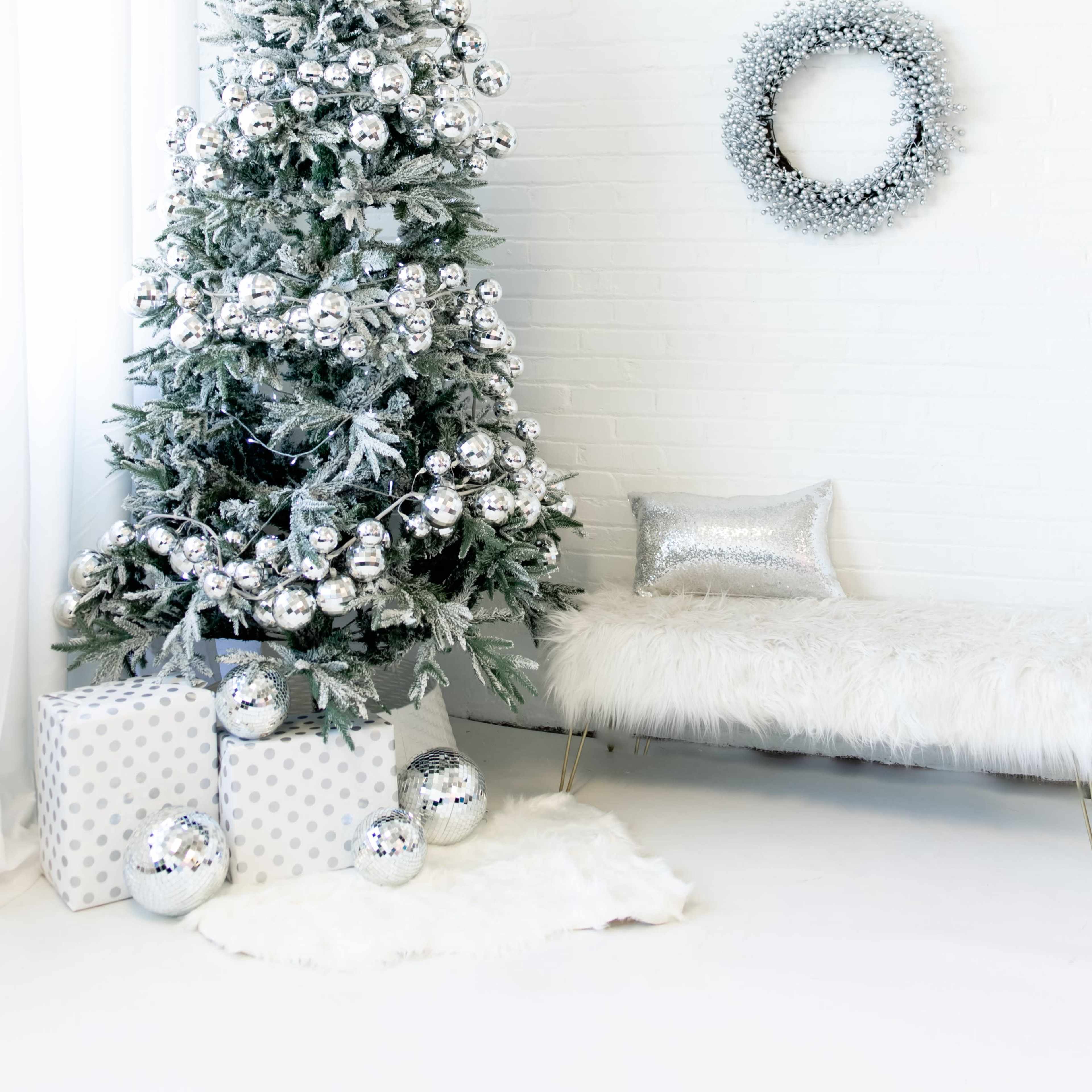 A decorated Christmas tree with silver ornaments stands next to a white bench and wrapped presents in a bright, minimalistic room.