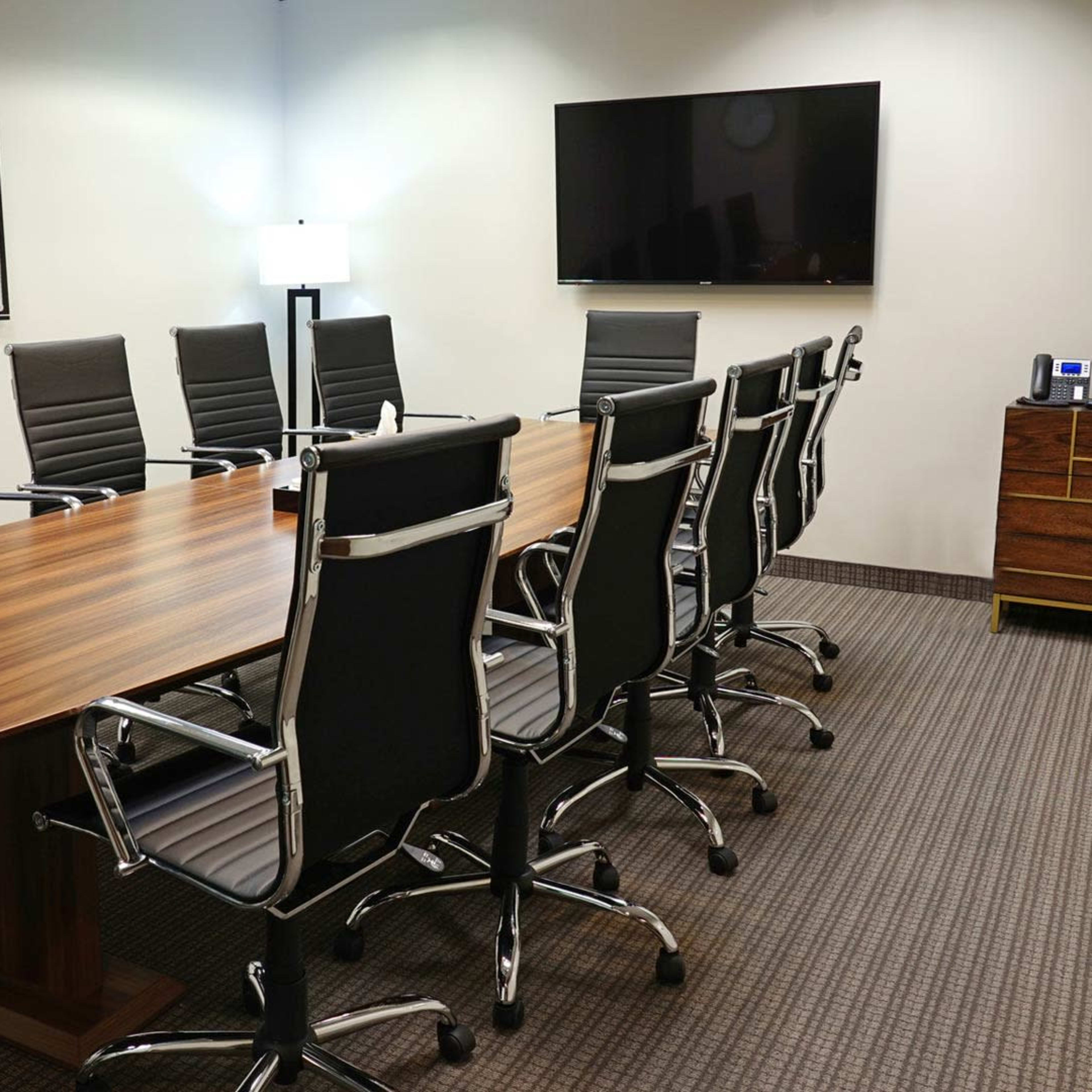 A modern conference room with a long wooden table surrounded by black ergonomic chairs, and equipped with a wall-mounted television and a coffee machine on a side cabinet.