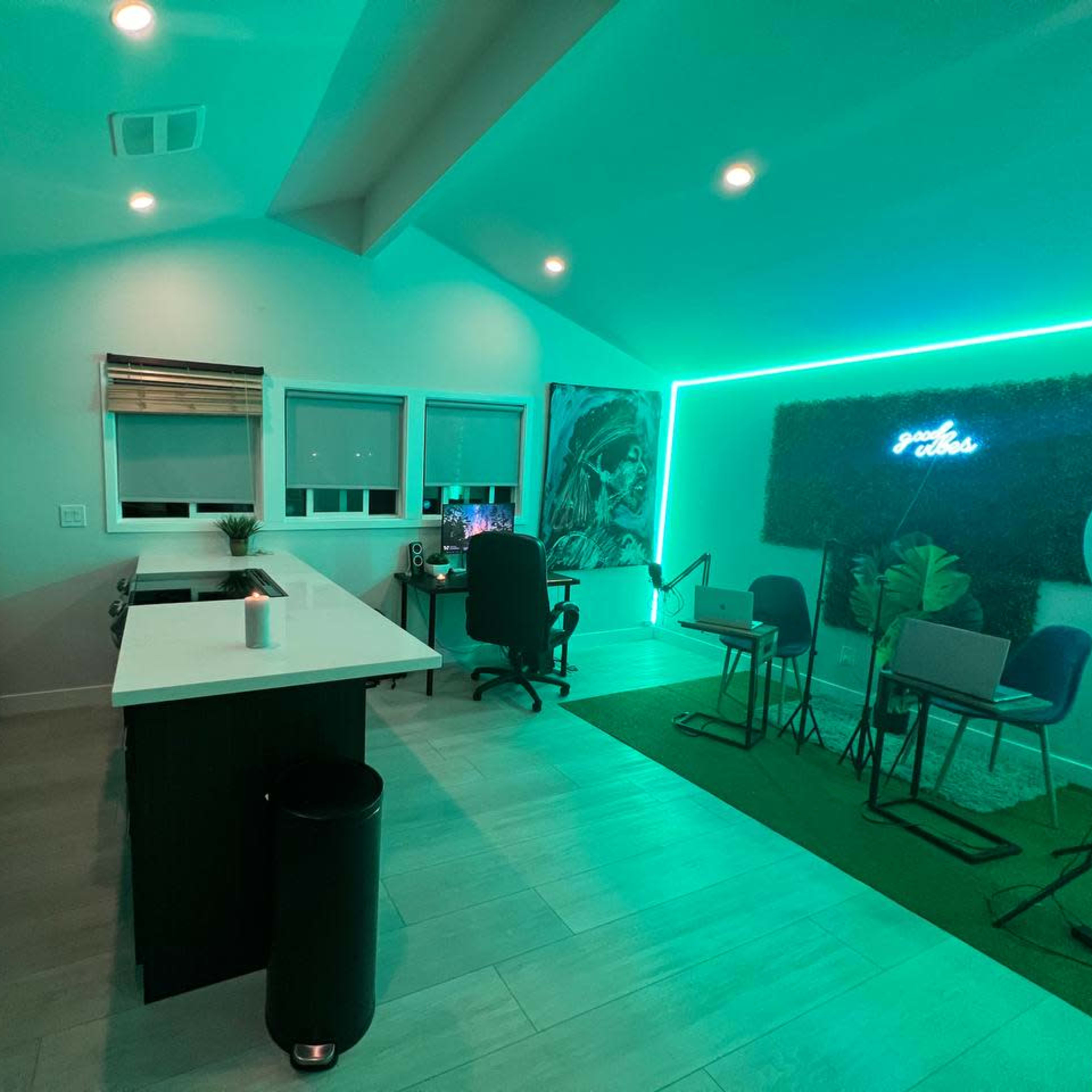 A modern kitchen and workspace area illuminated with green LED lighting, featuring a kitchen island, a desk with a computer chair, and a small seating arrangement.