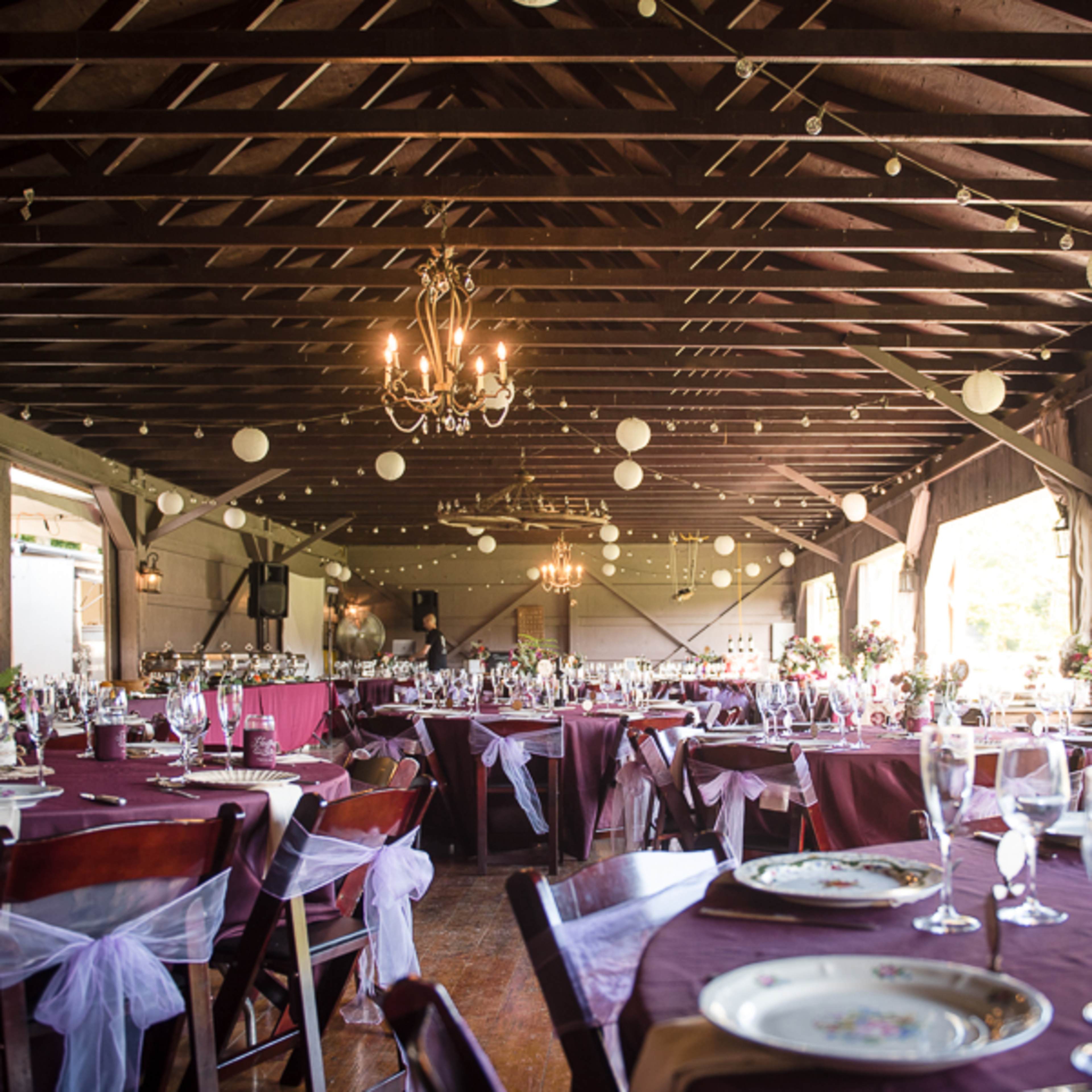 A spacious venue features several tables set with elegant tableware and decorations under a wooden beam ceiling.