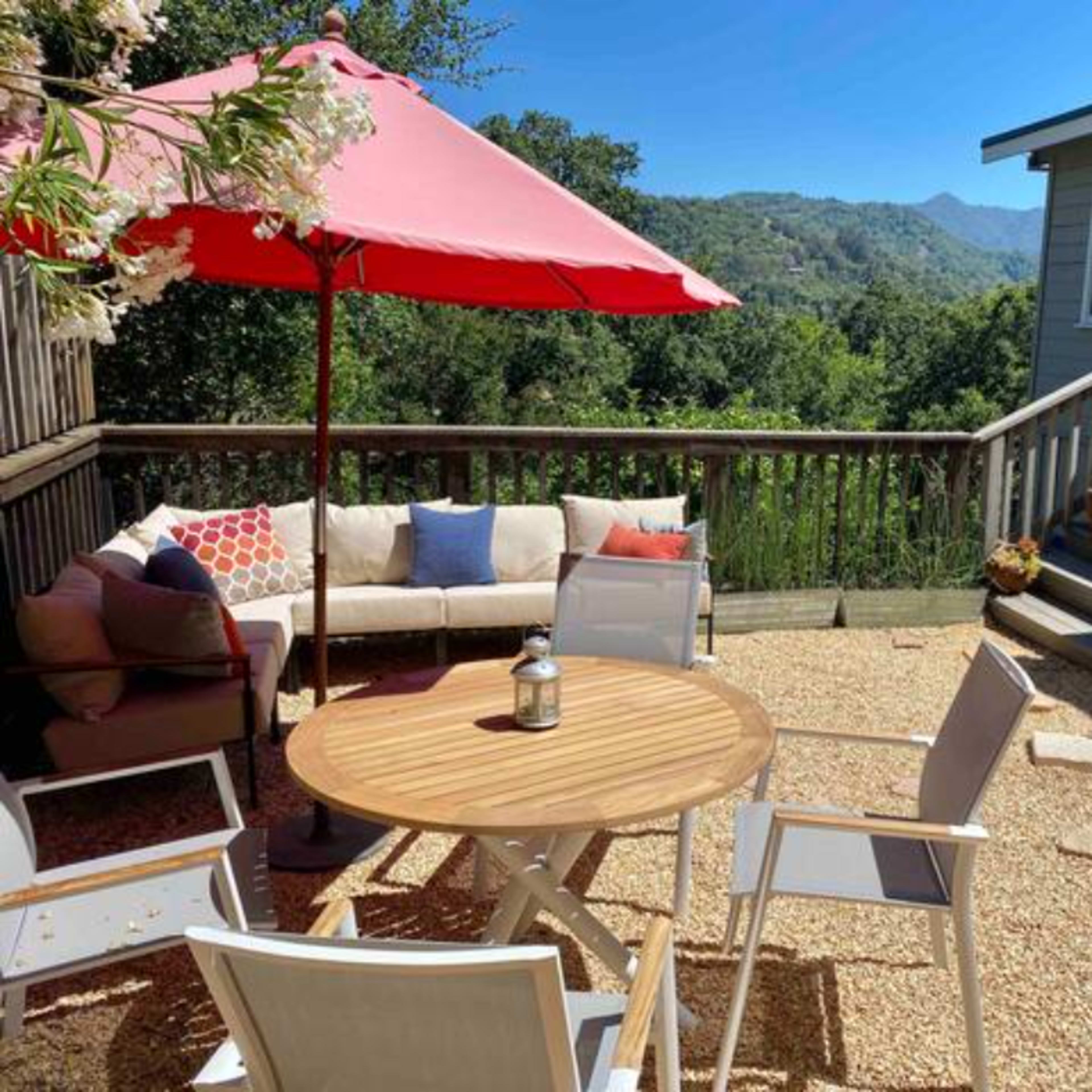 A patio features a round wooden table surrounded by white chairs, with a large red umbrella, a sofa, and a view of the hills in the background.