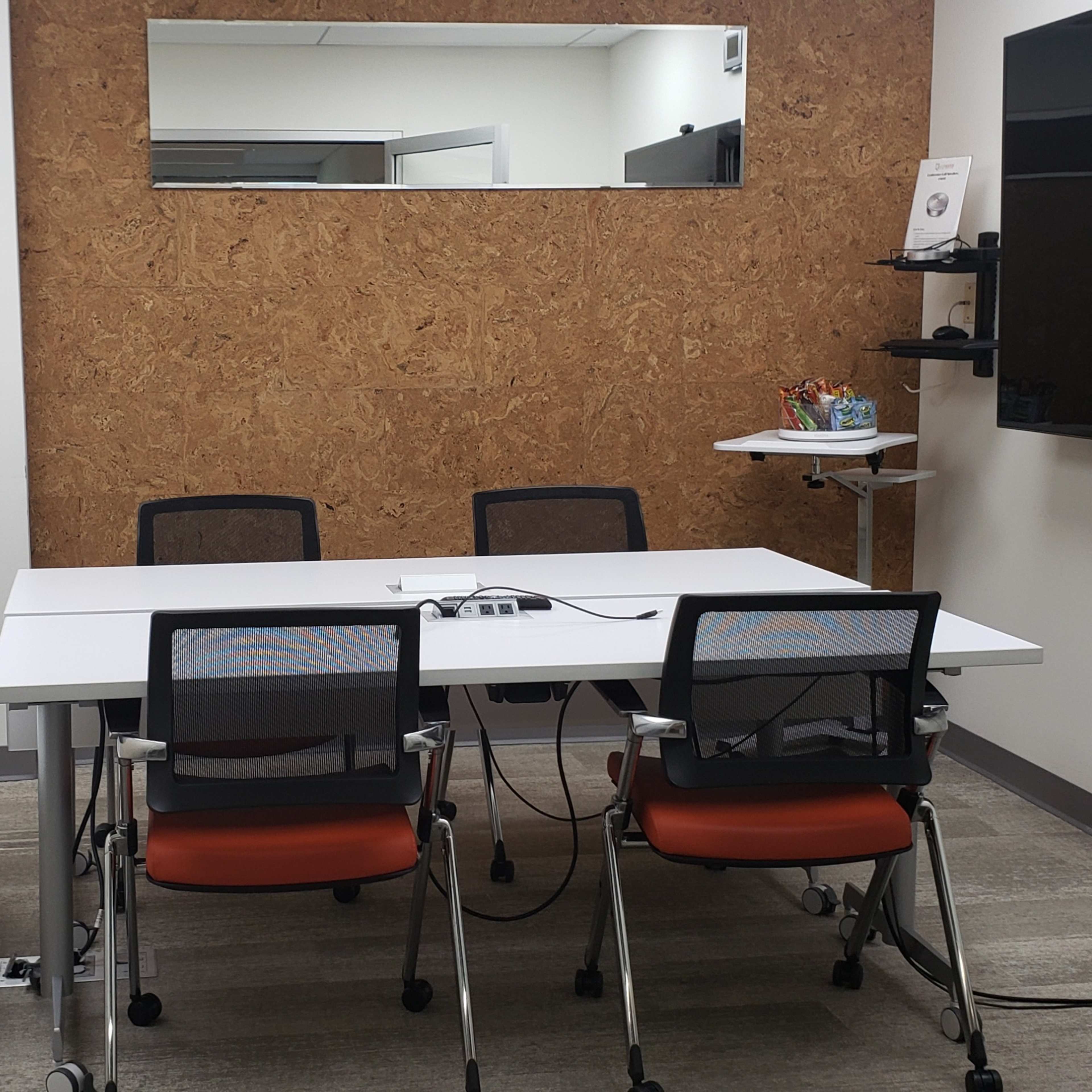 A small meeting room with a white table, four chairs, a wall-mounted screen, and a corkboard accent wall.
