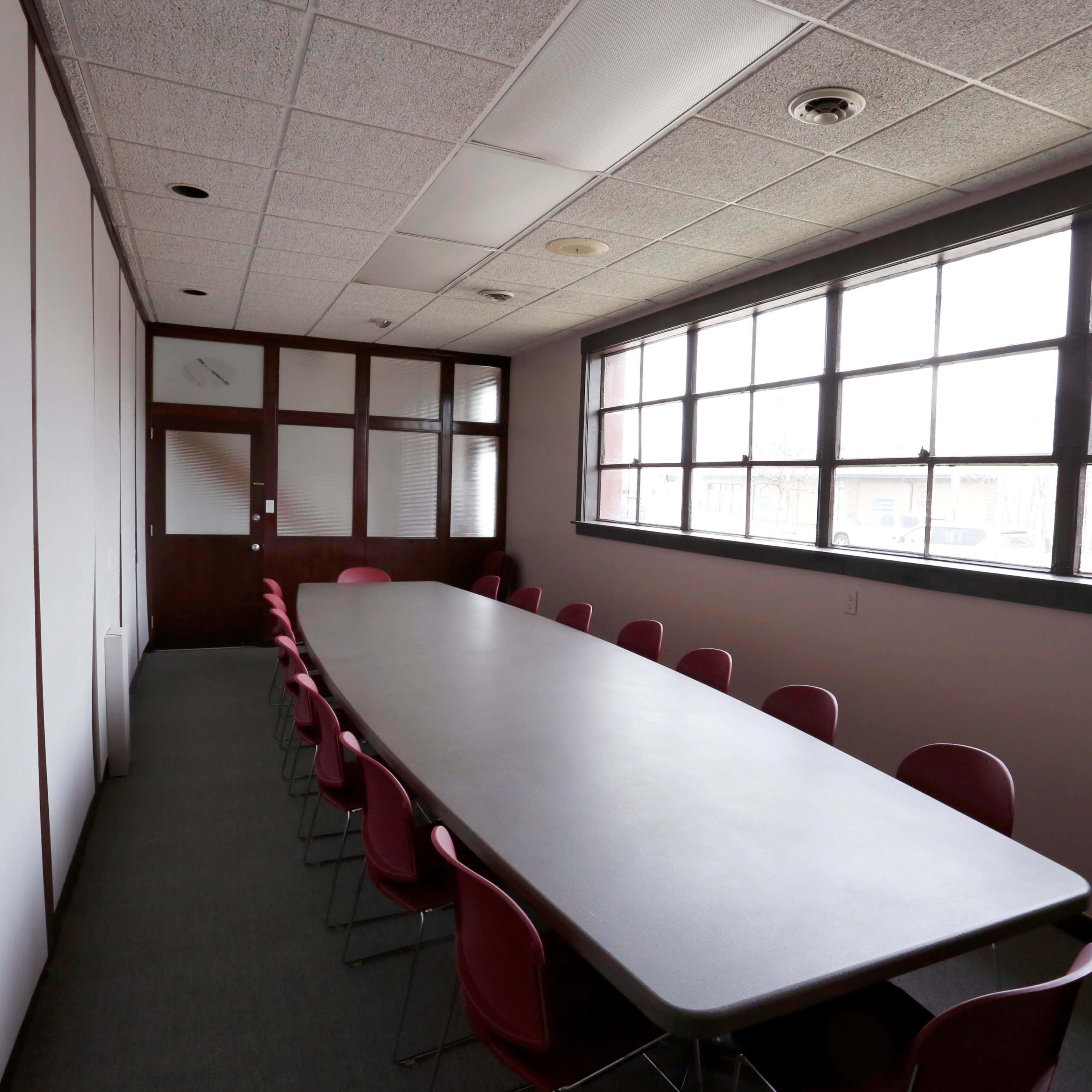A long conference table with red chairs is positioned in a sparsely furnished meeting room with large windows.