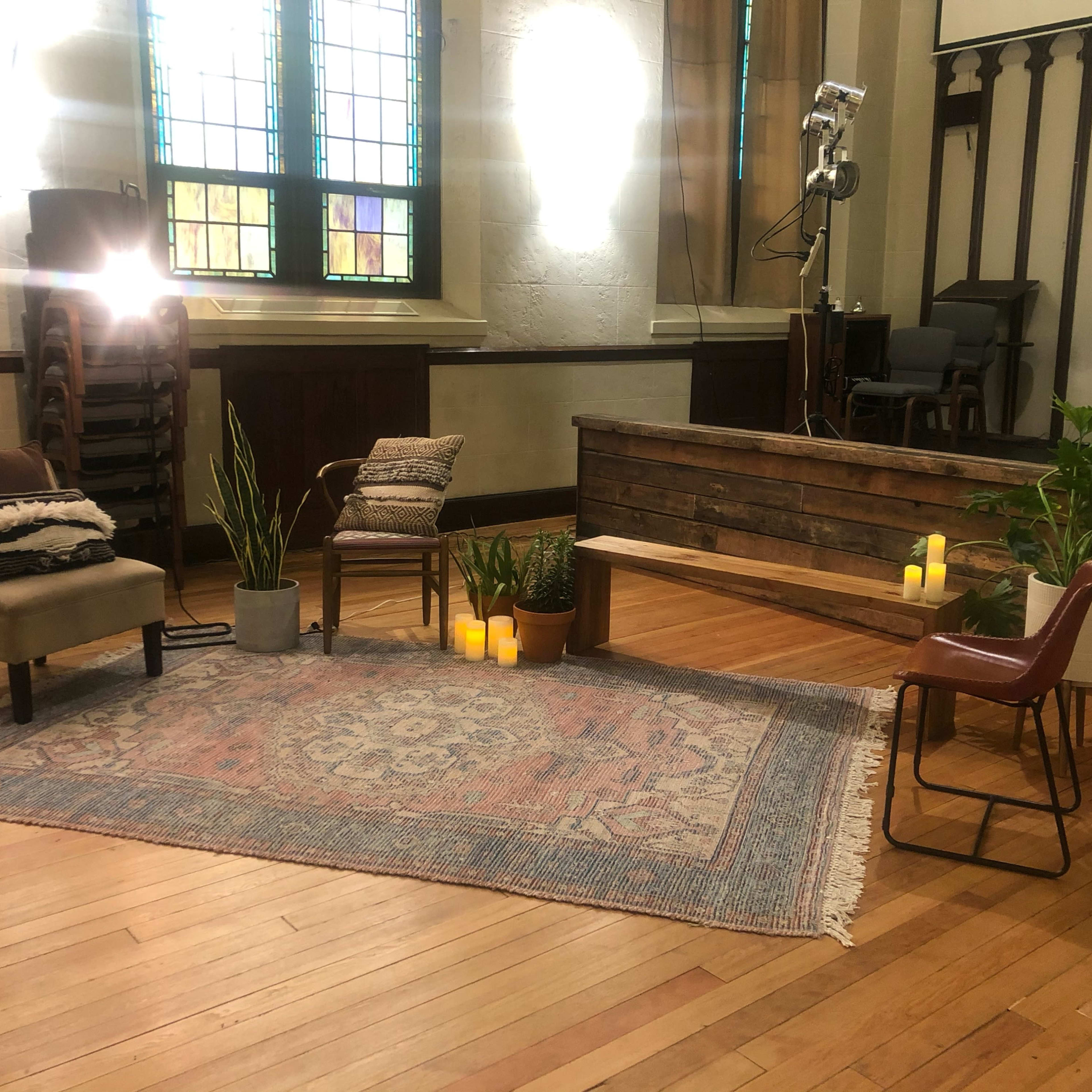 A cozy interior space with a wooden bench, two chairs, potted plants, a patterned rug, and candles arranged in a welcoming layout.