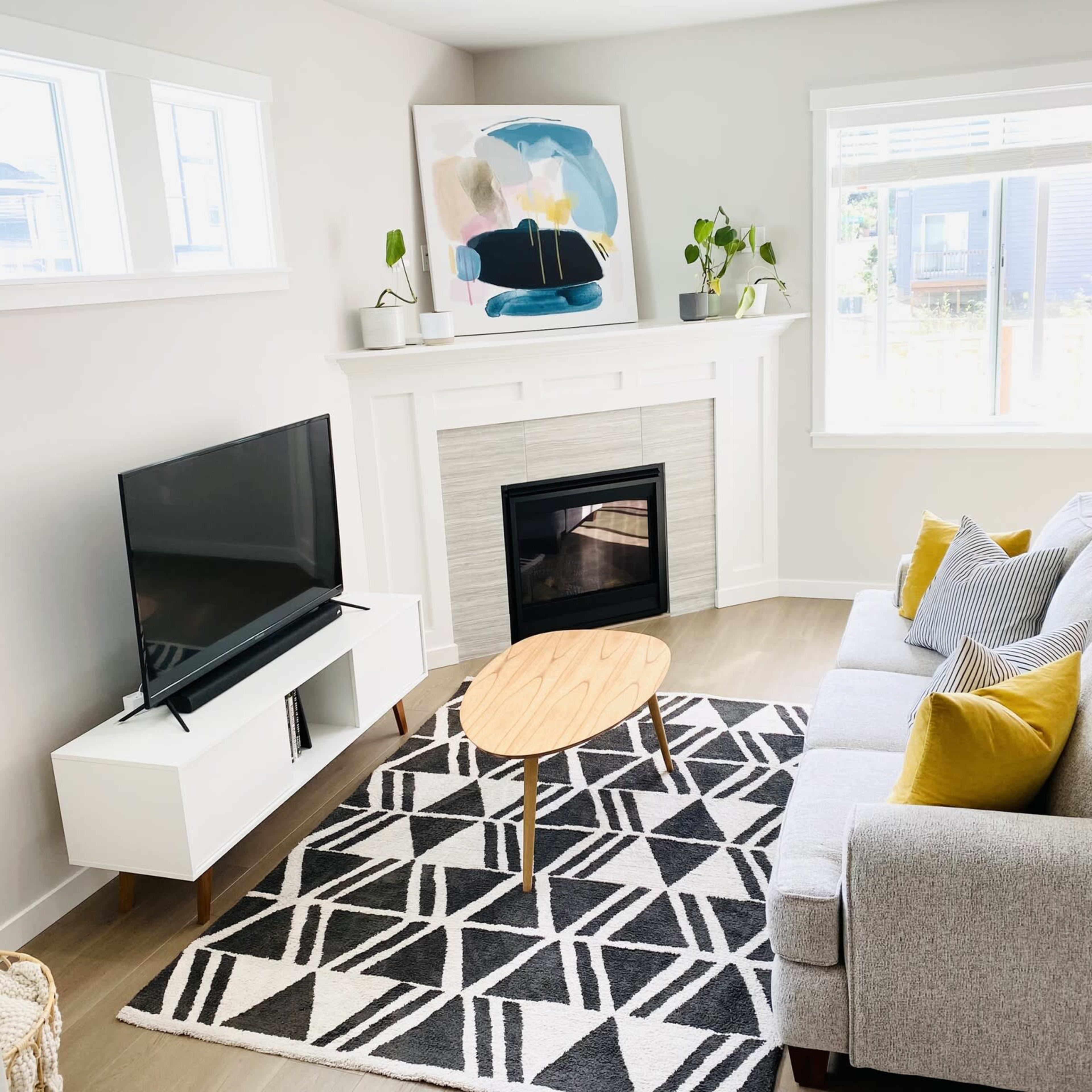 A modern living room features a gray sofa, a wooden coffee table, a television on a white media console, and a fireplace beneath a large artwork, with natural light coming through the windows.