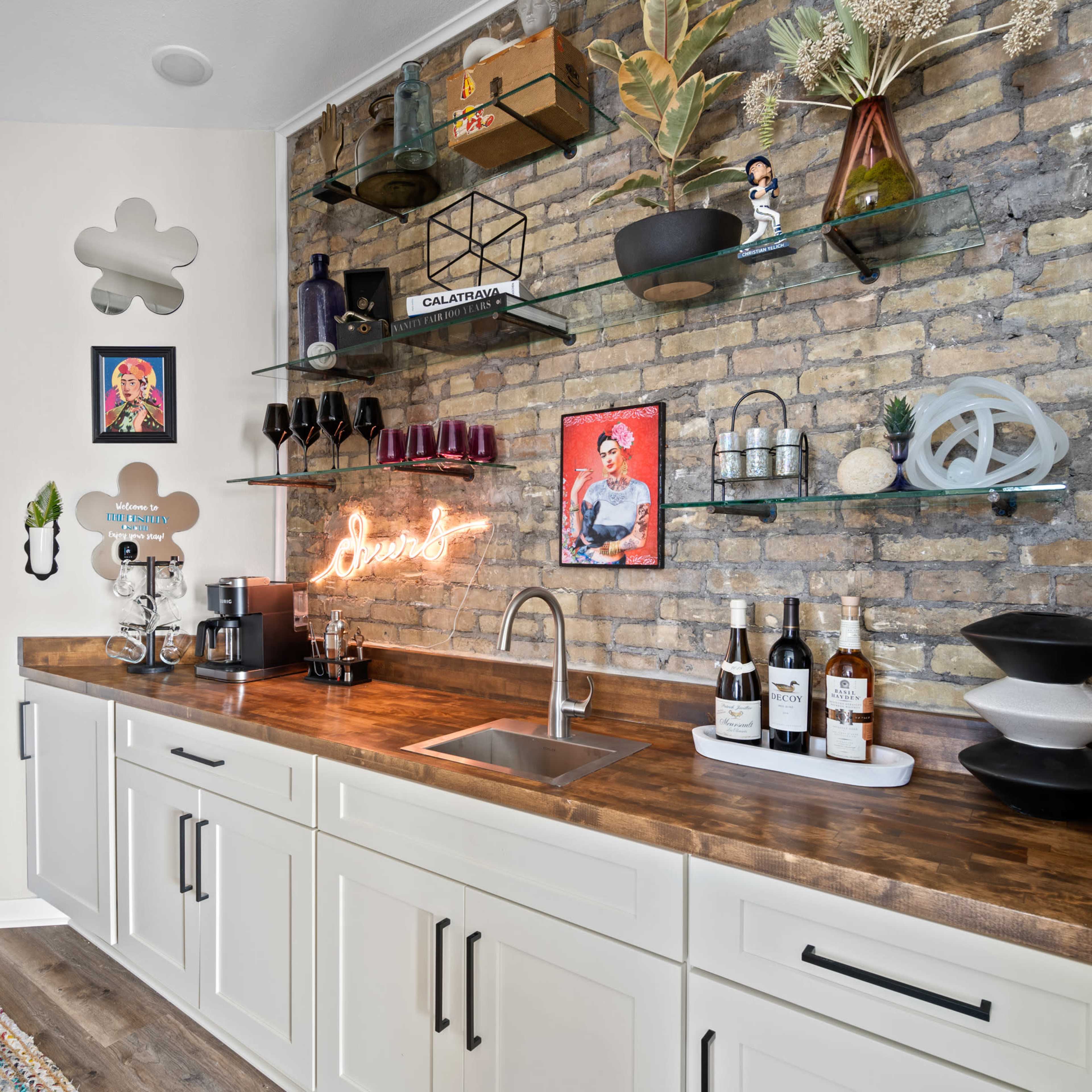 A modern kitchen bar area featuring a wooden countertop, open shelving with decorative items, a sink, and various bottles of wine displayed.
