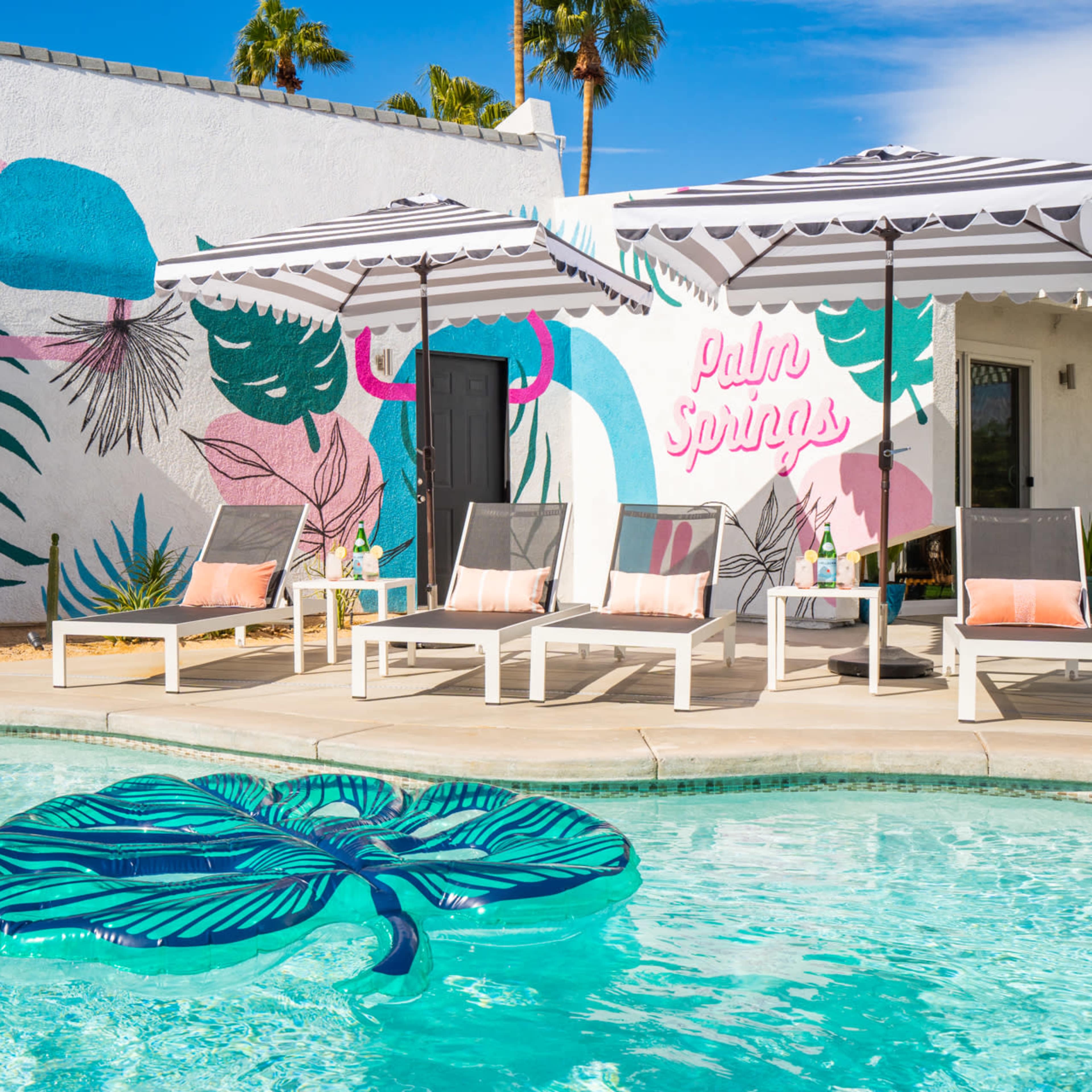 A vibrant pool area with lounge chairs and a large leaf-shaped float, set against a colorful mural and palm trees.