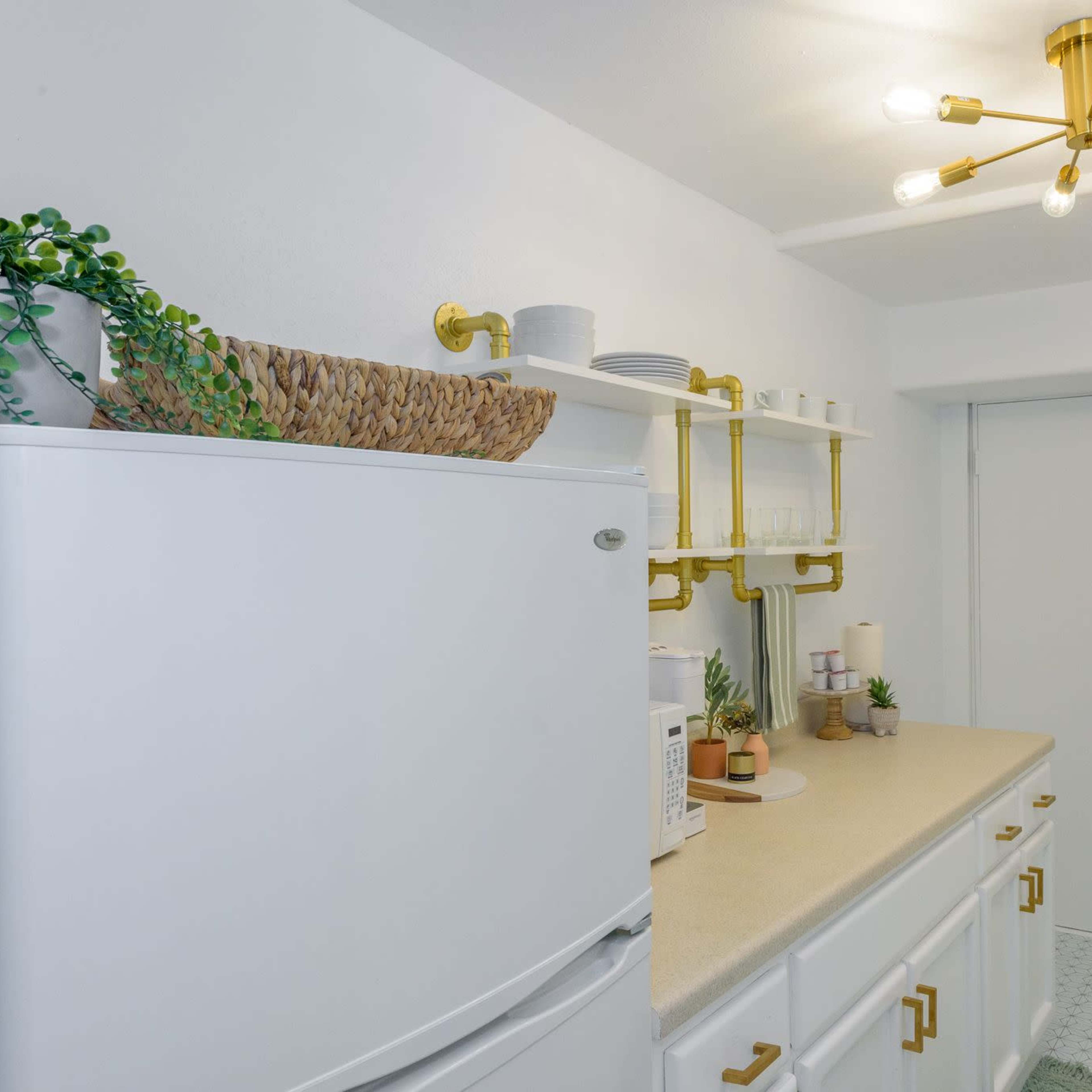 A modern kitchen with a white refrigerator, a countertop featuring a microwave and plants, and decorative shelves above with dishware and a framed plant artwork on the wall.