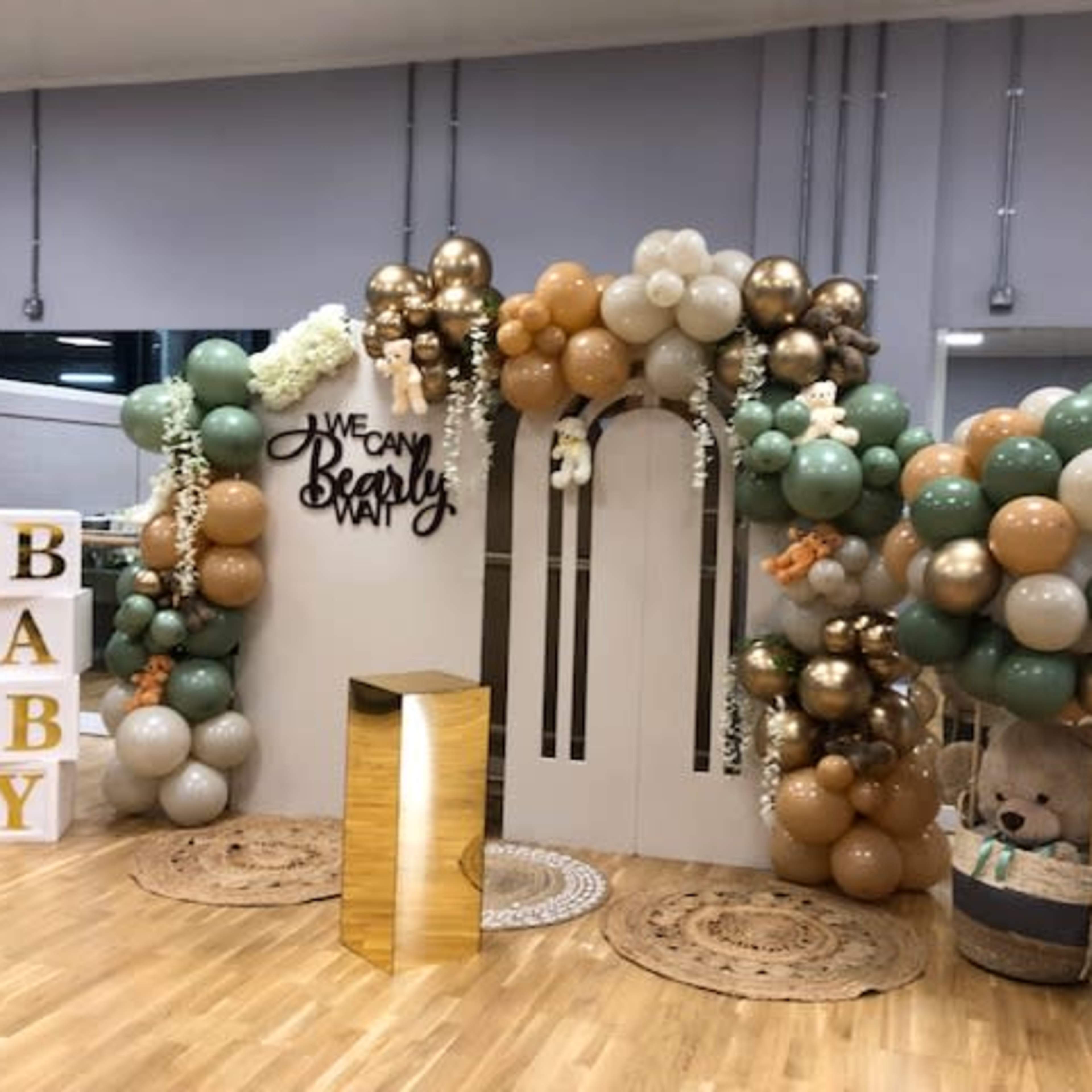 A festive decoration setup featuring a balloon arch in various colors, a gold pedestal, and thematic signage that reads "WE CAN BEARLY WAIT" alongside a large "BABY" display.
