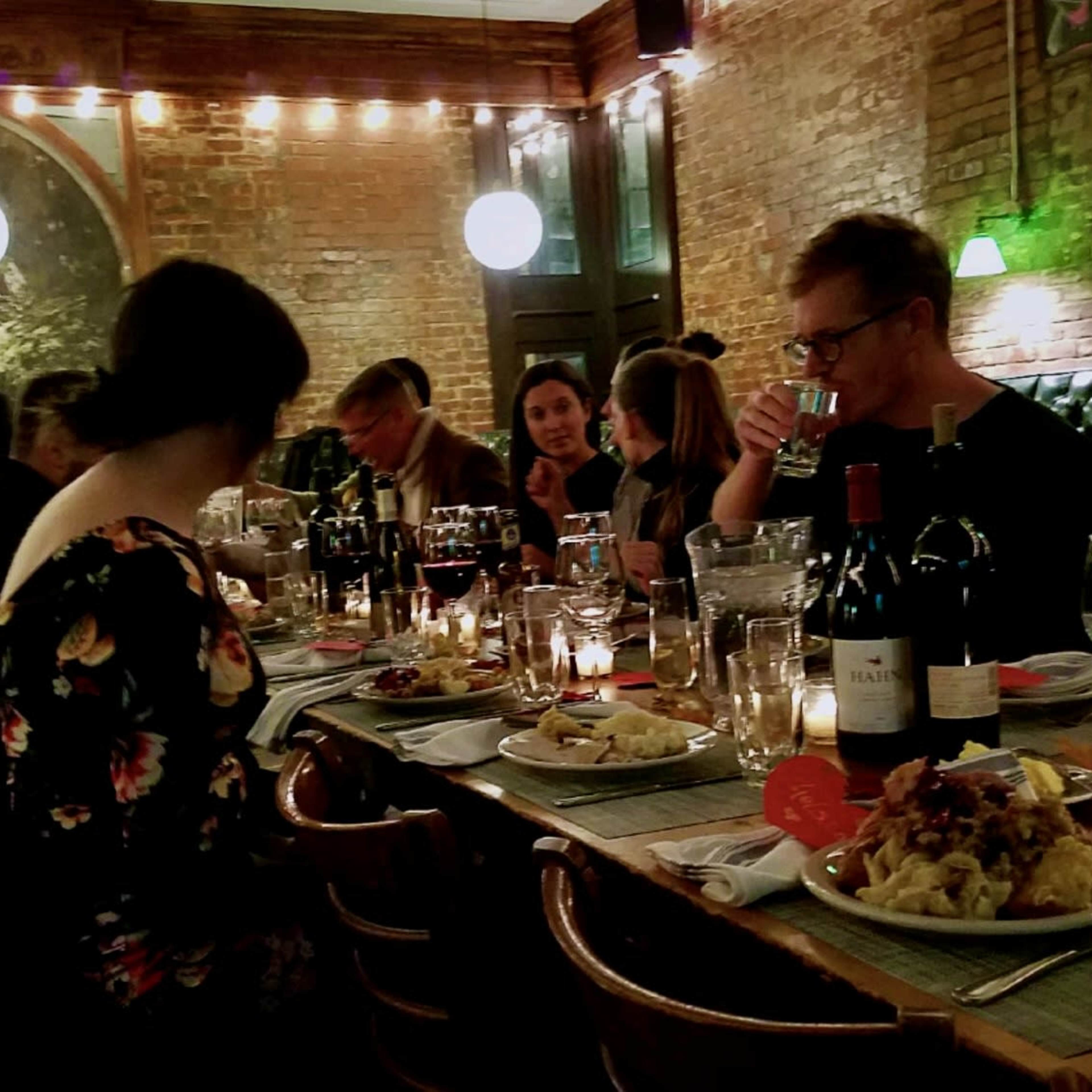 A group of people sits around a long table filled with food and wine in a warmly lit restaurant.