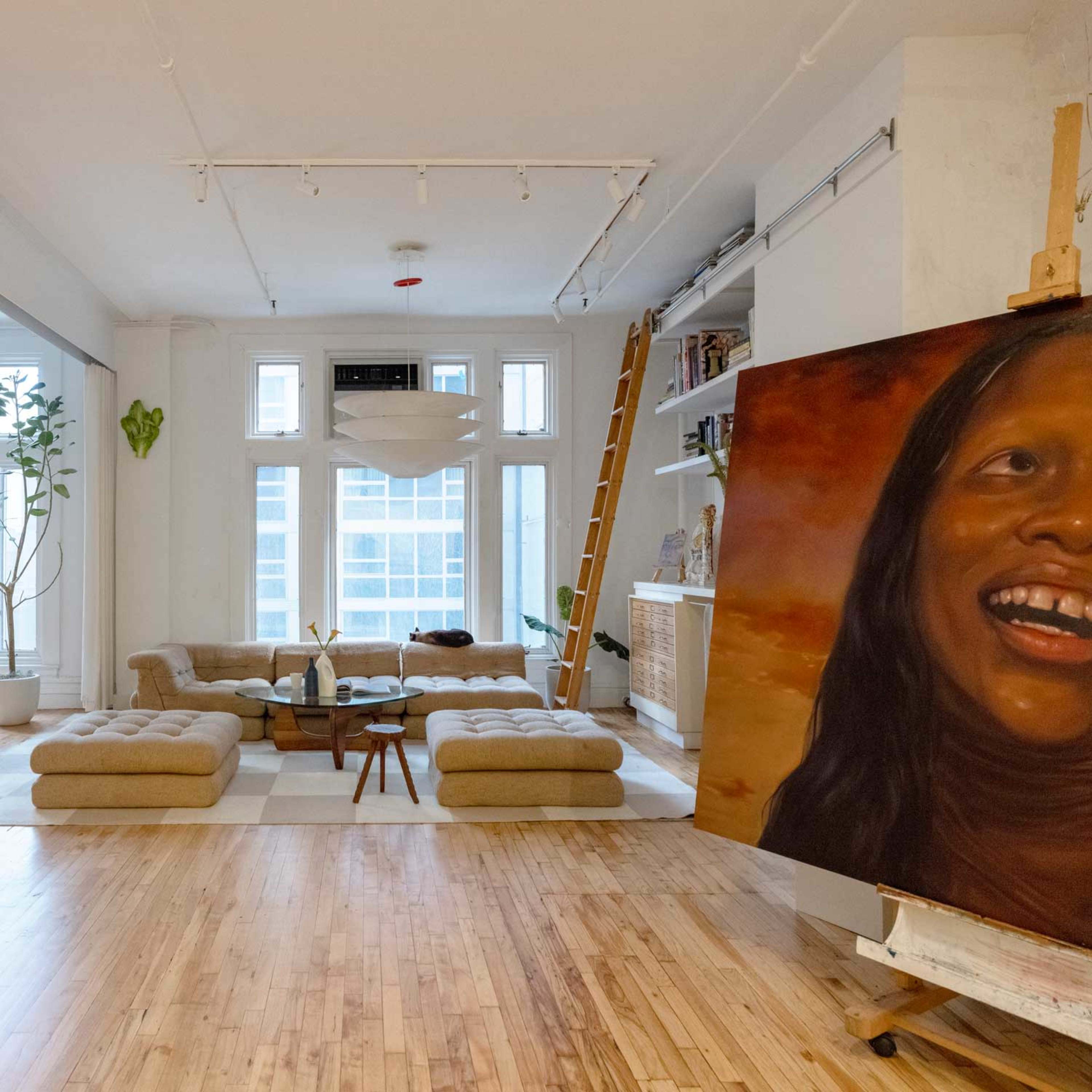 A spacious, modern living room with a large painting on an easel, a sofa set in the center, and large windows letting in natural light.