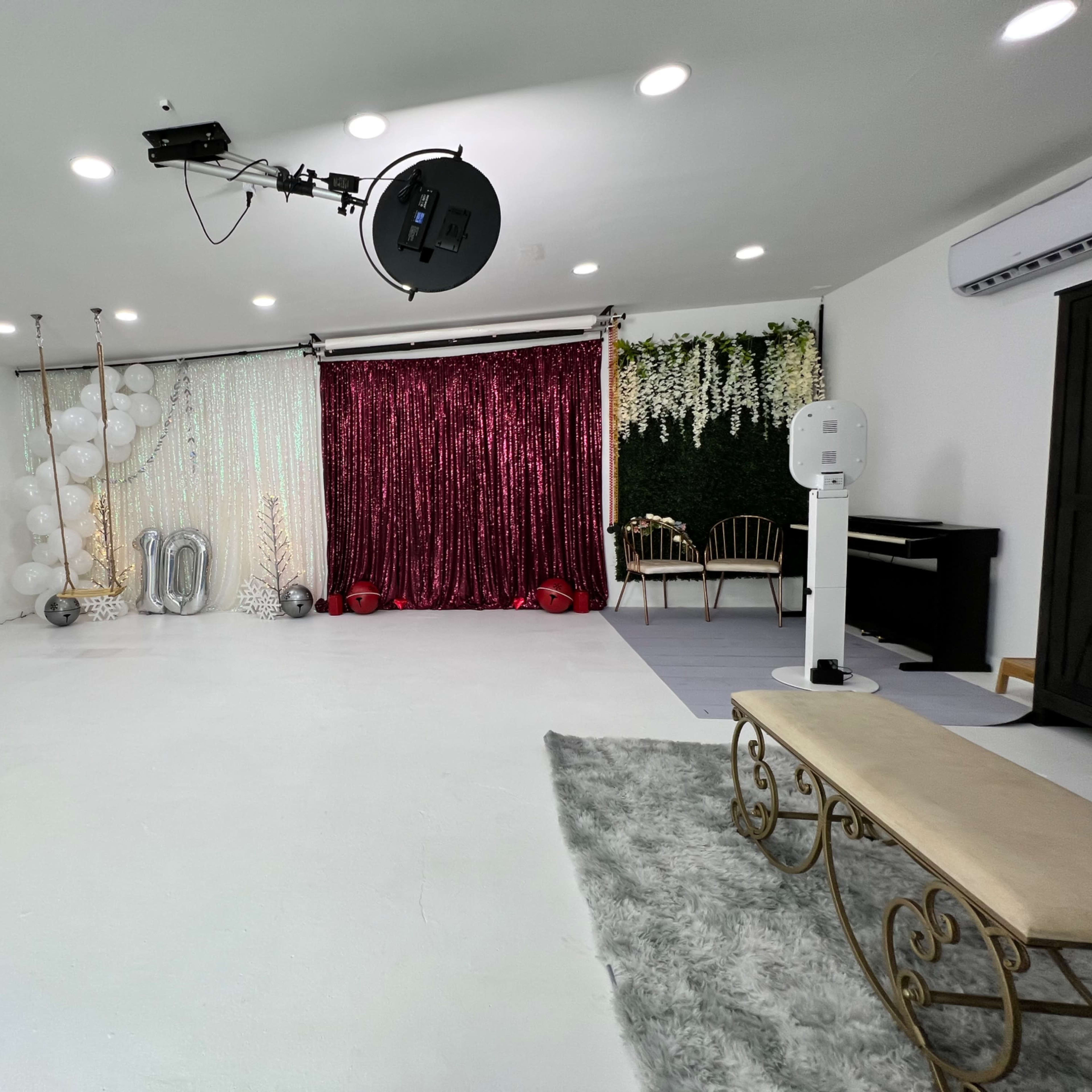 A well-decorated studio space with a white floor, a bench, greenery, balloons, a glittery backdrop, and a piano.