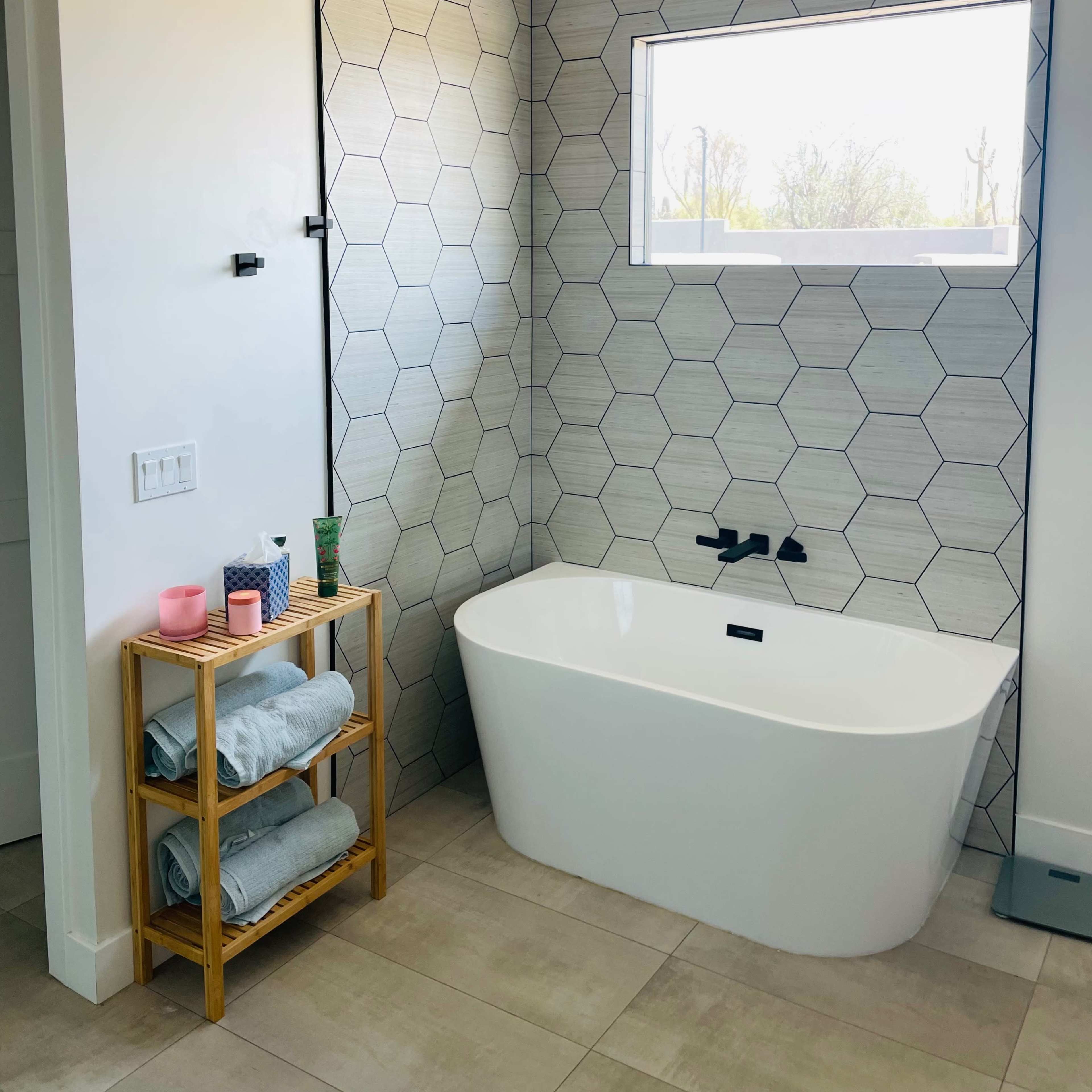 A modern bathroom with a hexagonal tiled wall, a freestanding bathtub, and a bamboo shelf holding towels and toiletries.