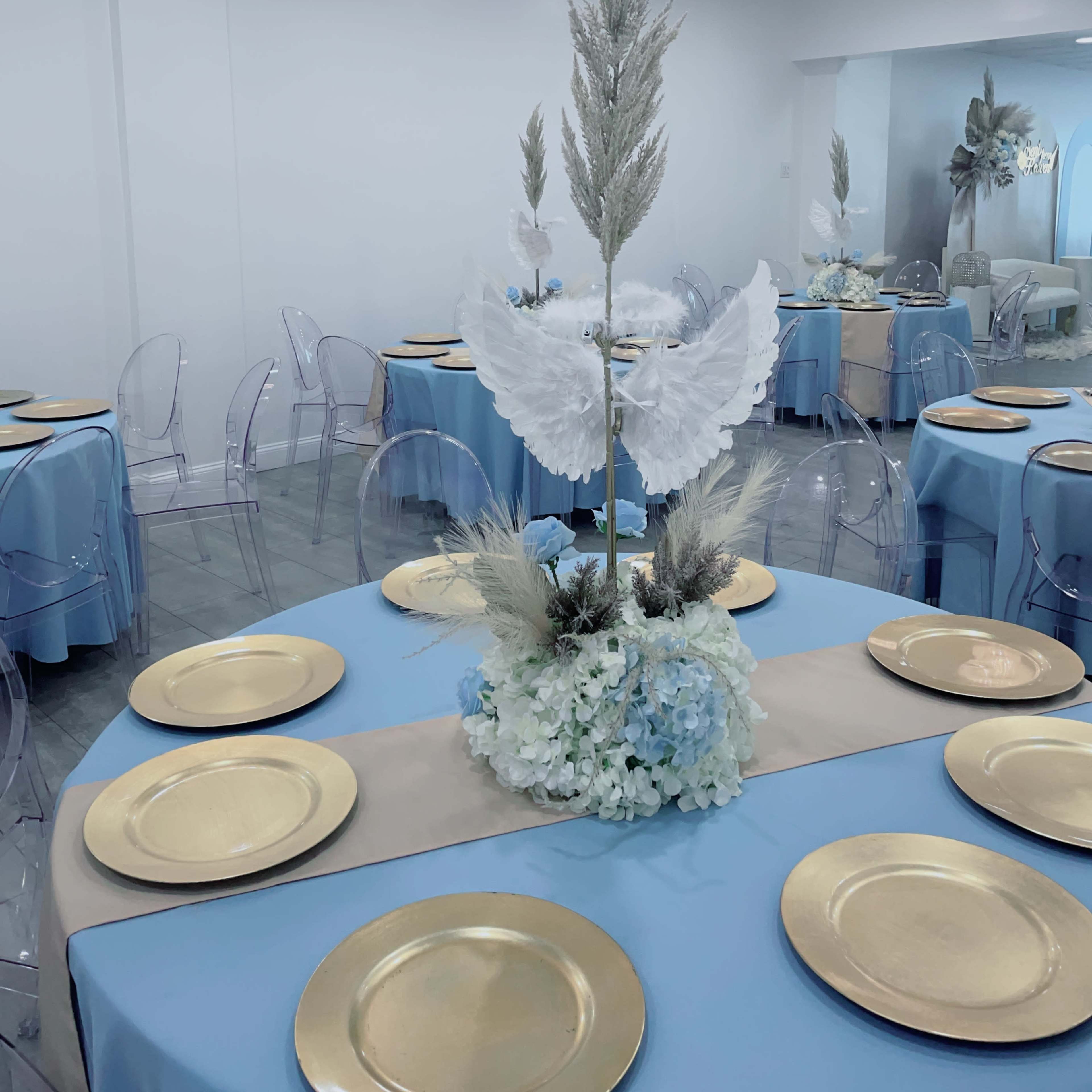 A dining area is set with tables dressed in blue and beige linens, featuring gold plates and decorative centerpieces of white and blue flowers.