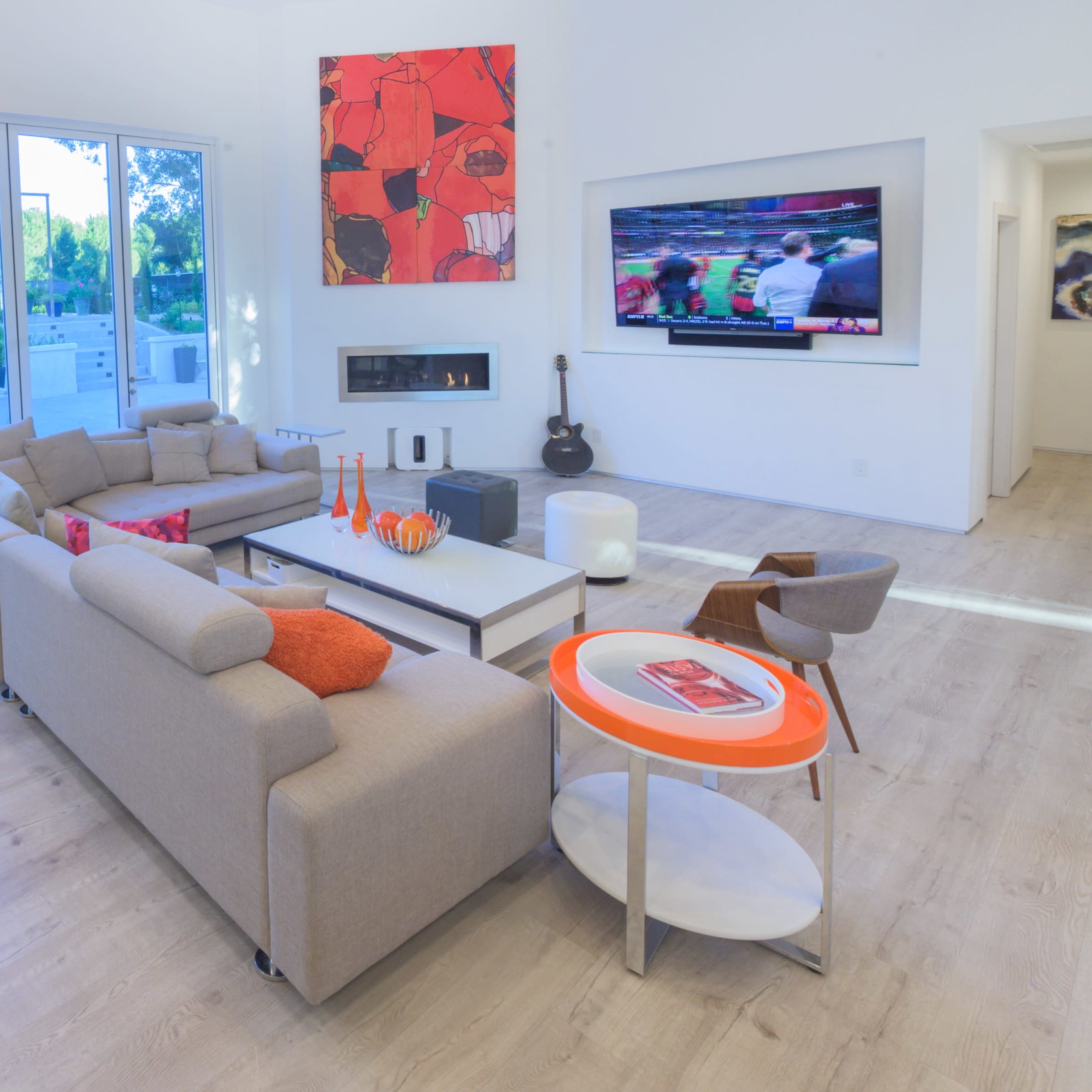 A modern living room features a beige sectional sofa, a television mounted on the wall, and large windows overlooking an outdoor space.