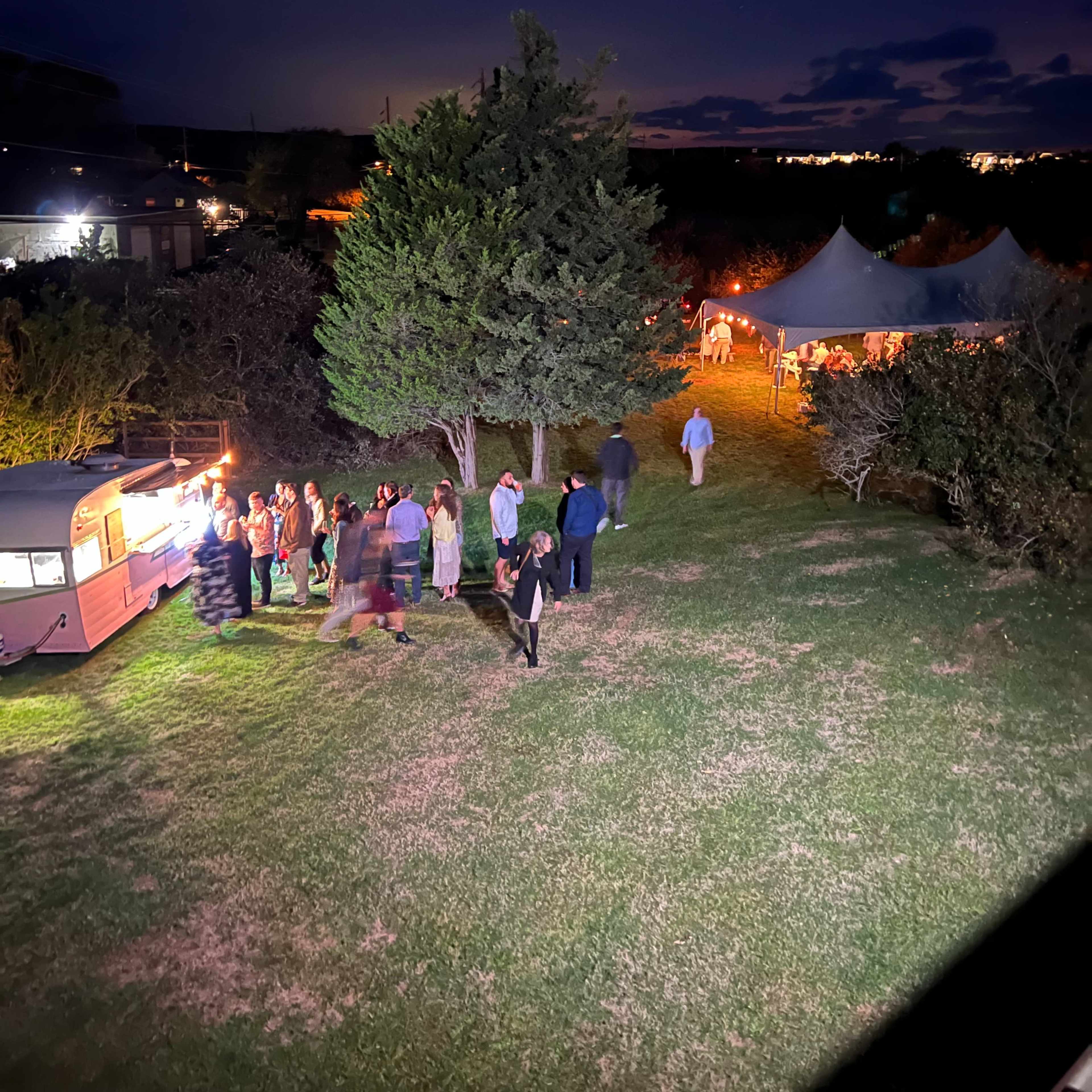 A group of people lines up at a food vendor near a tented event space under a twilight sky.