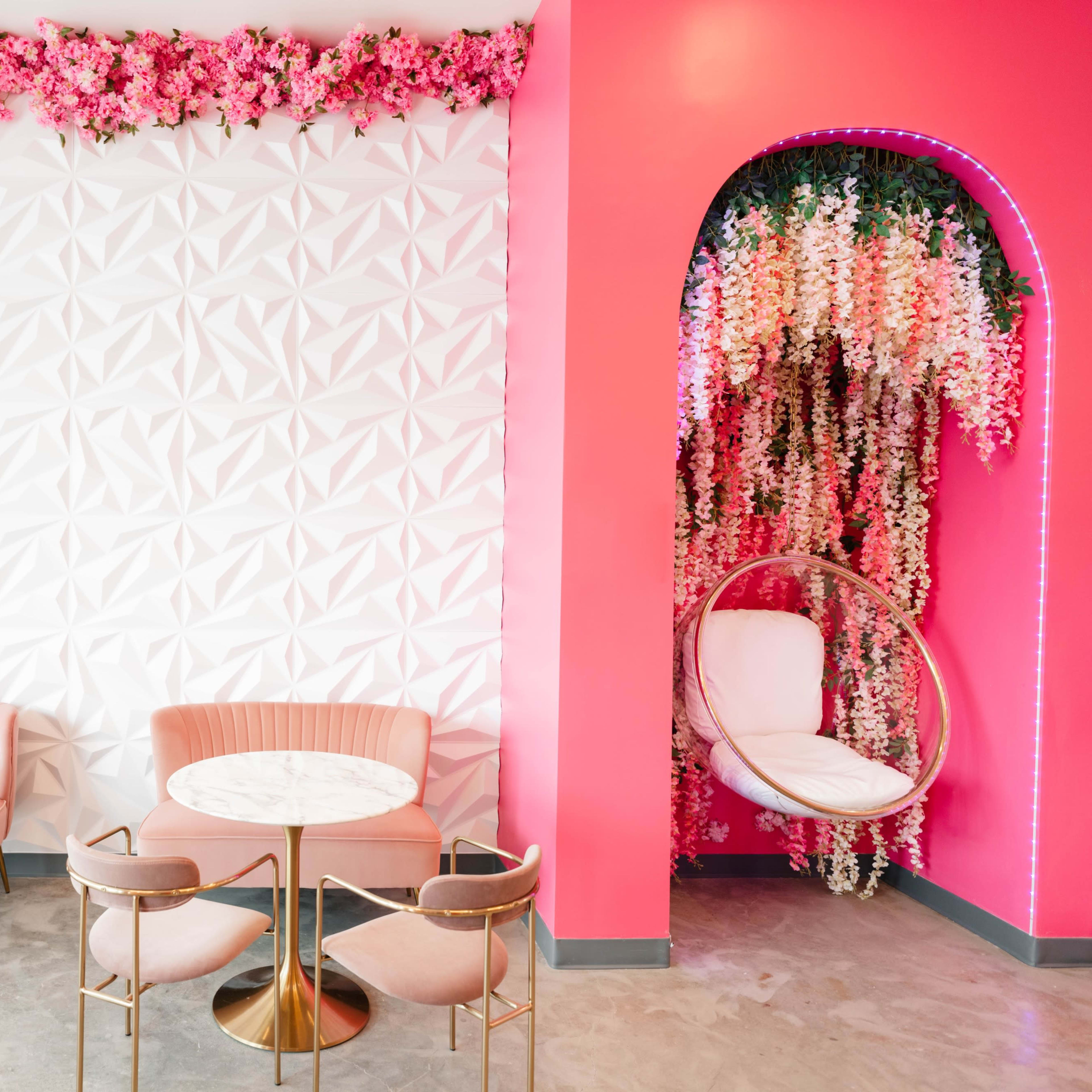 A stylish café interior featuring pink walls adorned with artificial flowers, marble-topped tables, and plush seating.