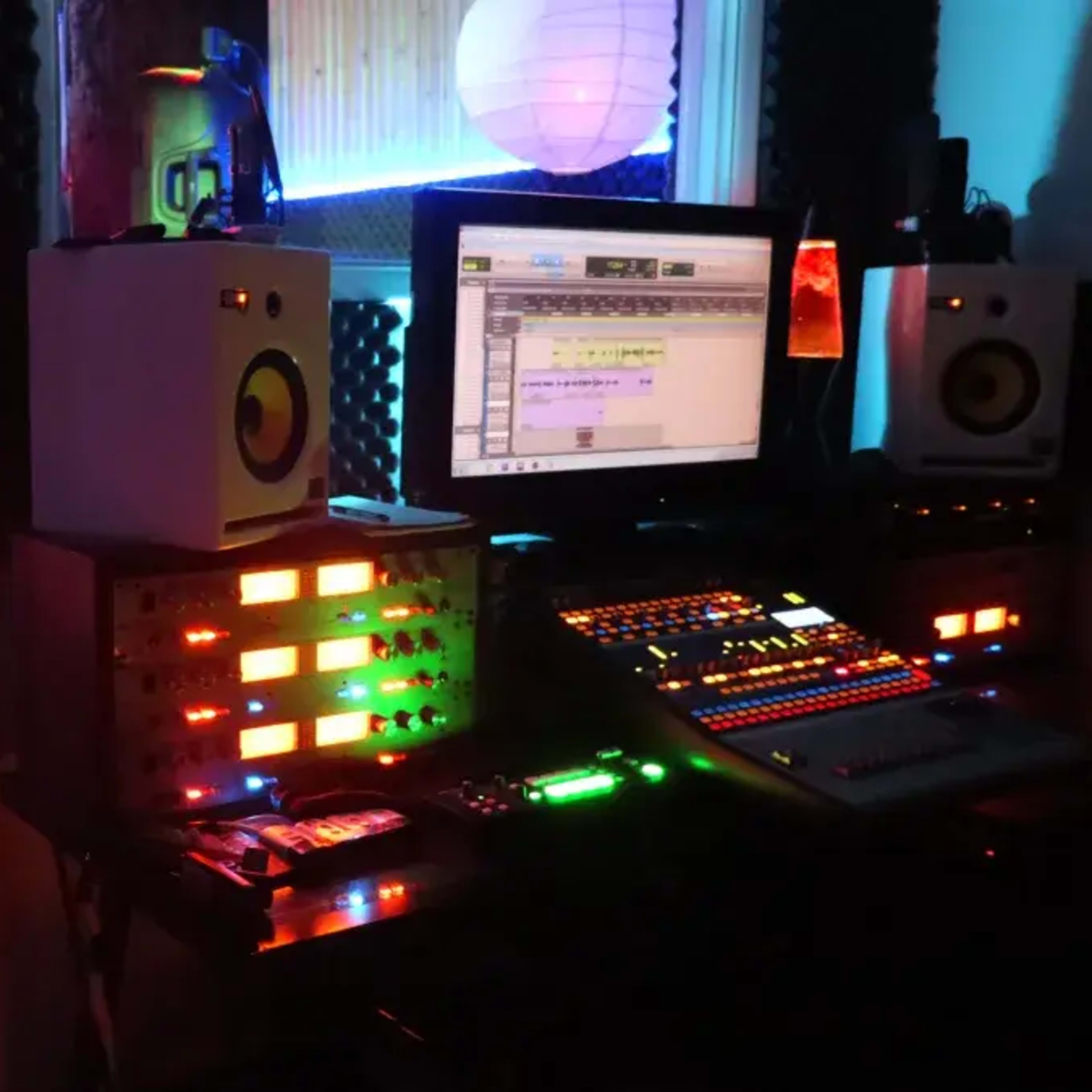 A music production studio with a control desk featuring colorful lights and a computer monitor, beside a lit performance area with a blank backdrop.