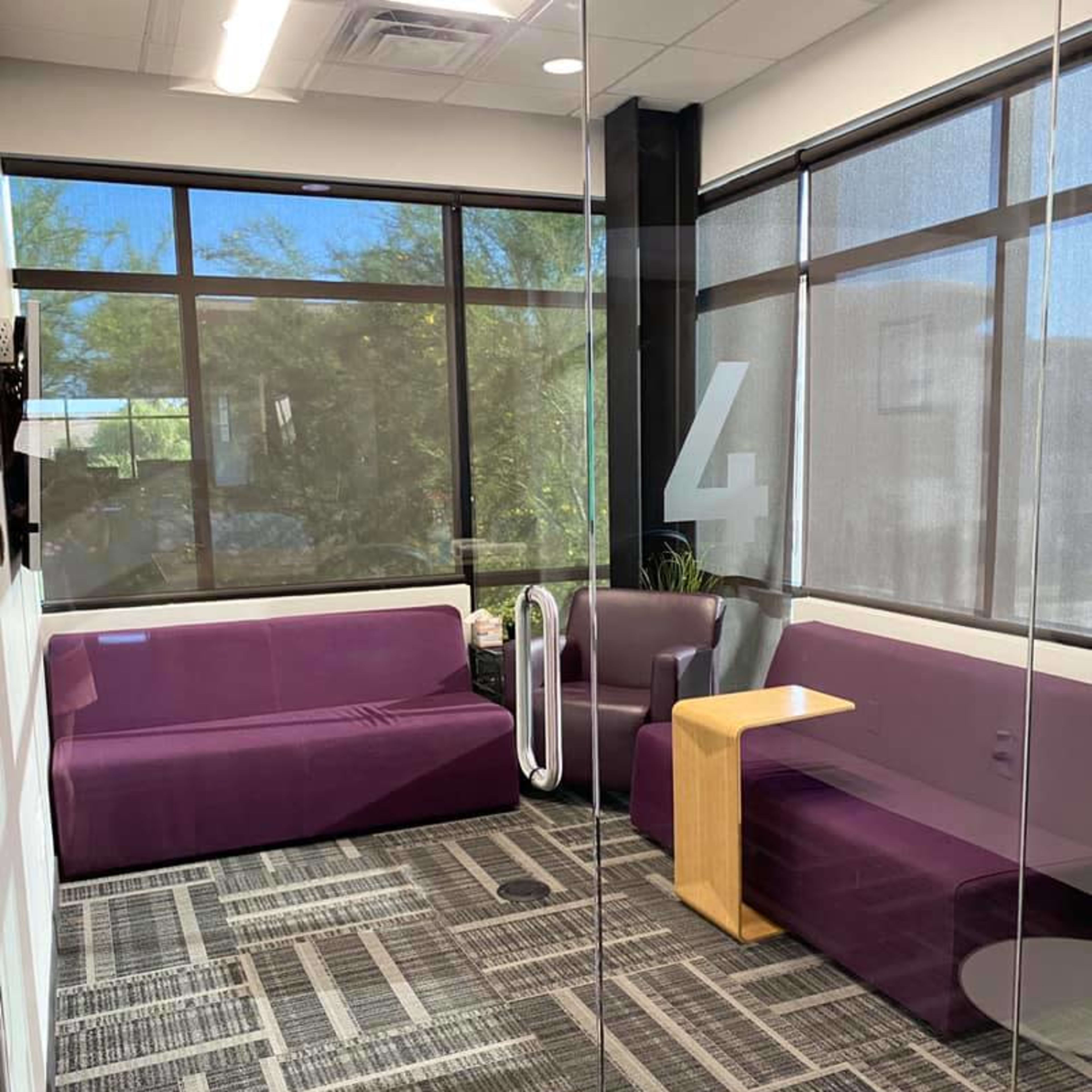 A modern office lounge area with purple seating, a wooden side table, and large windows overlooking greenery.