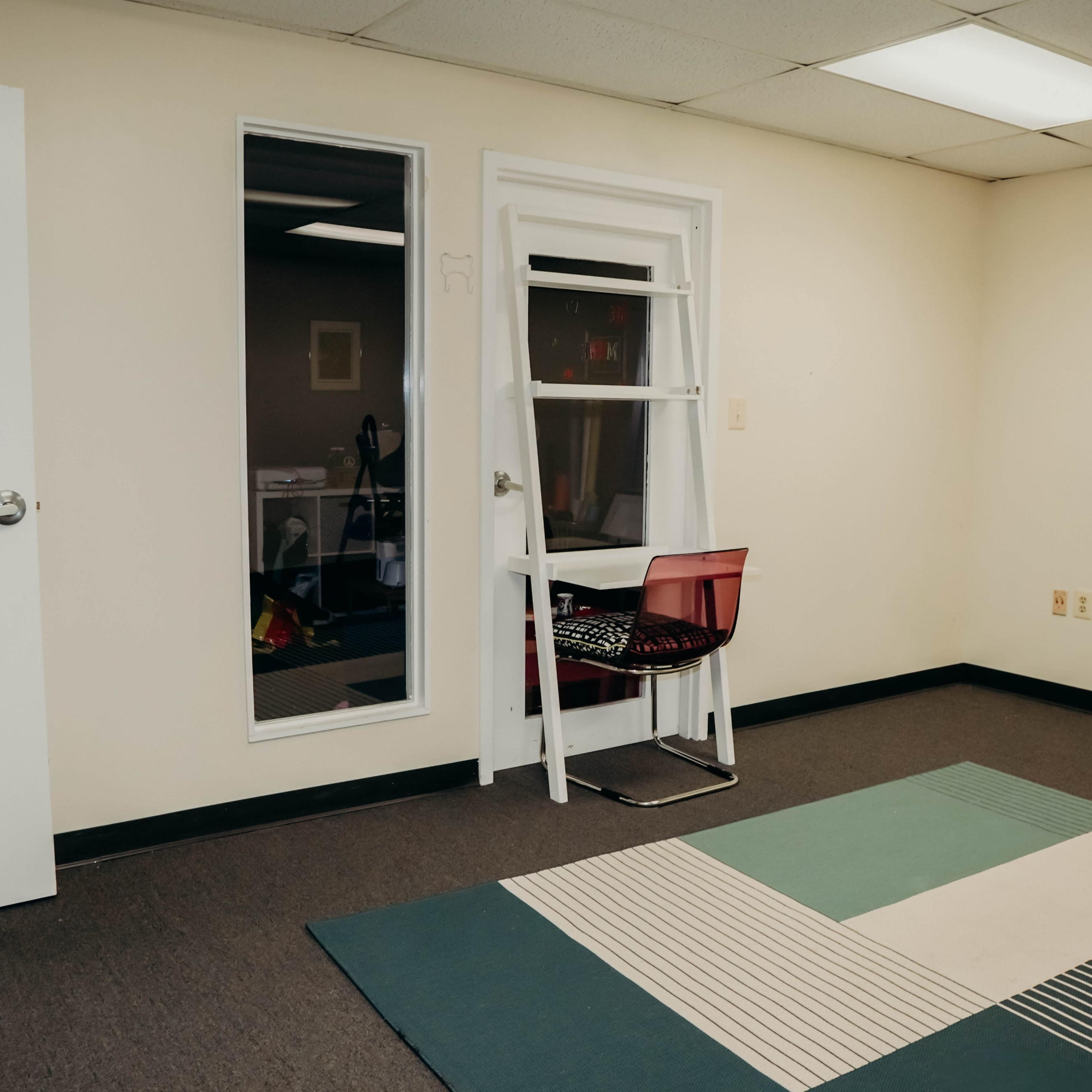A bare room features a chair positioned near a window and a large, multi-colored rug on the floor.