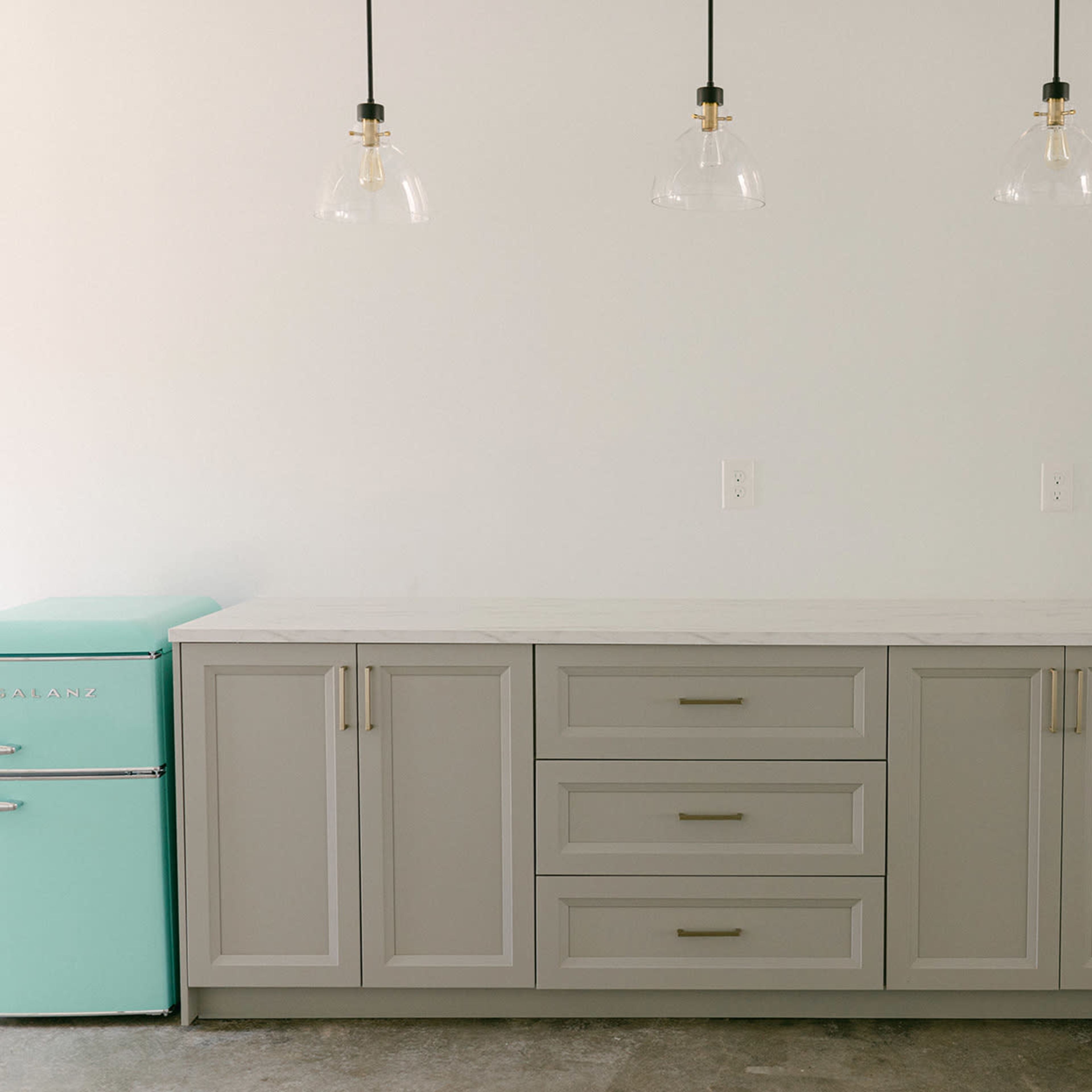 A kitchen setup features a light gray cabinet with three drawers and a matching countertop next to a mint green retro-style refrigerator, illuminated by three pendant lights.