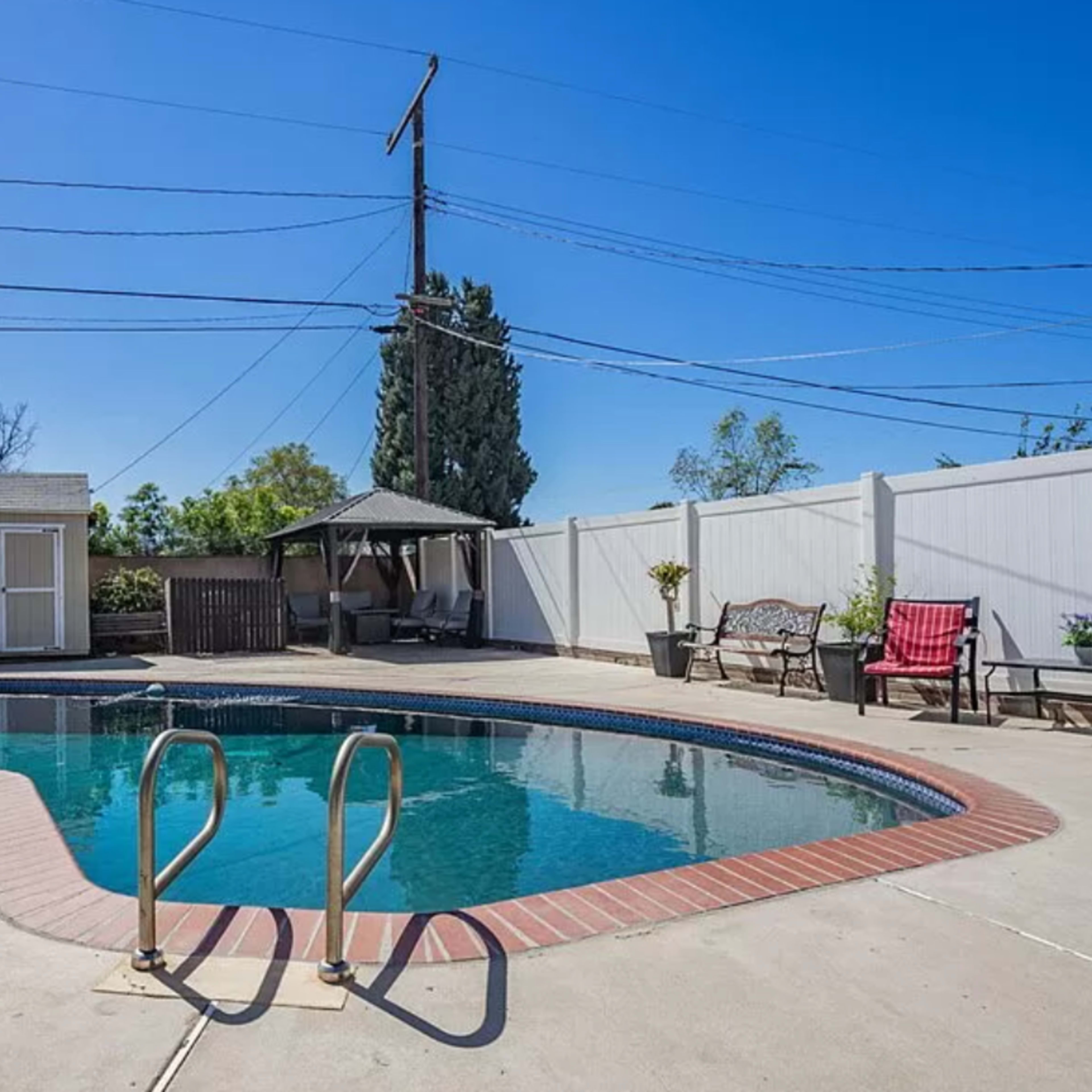 A backyard pool area with a rectangular swimming pool, a nearby shed, a gazebo, and seating arrangements along the perimeter.
