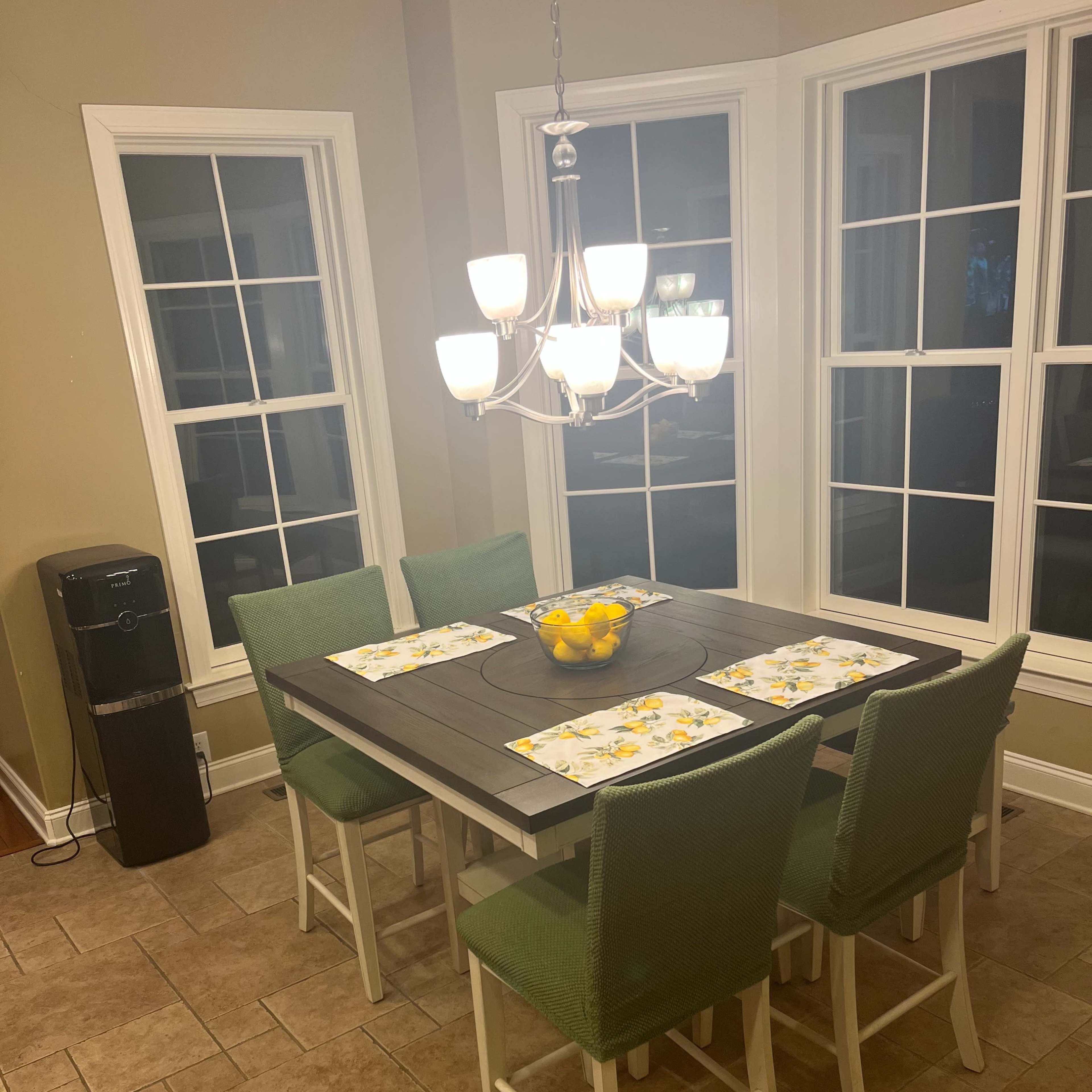 A wooden dining table with six green chairs is set with placemats and a bowl of lemons, surrounded by large windows.