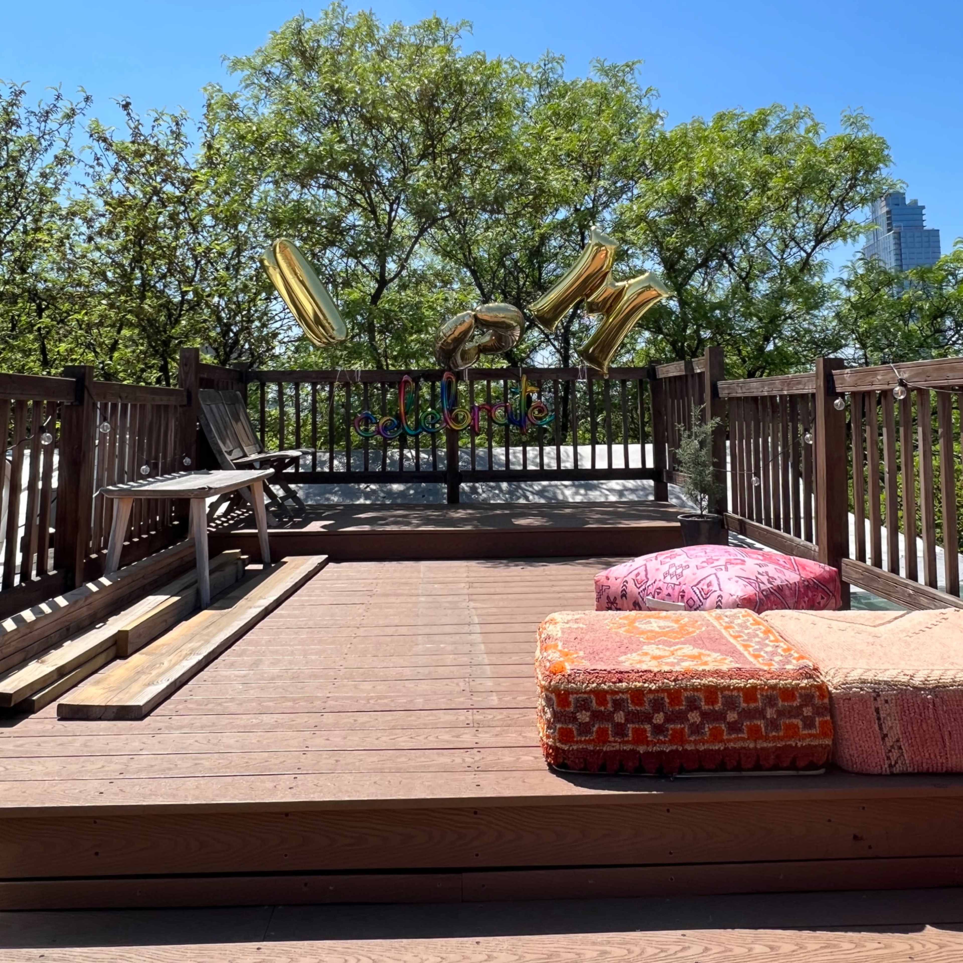 A wooden deck is decorated with colorful pillows and large gold balloons against a clear blue sky and lush green trees.