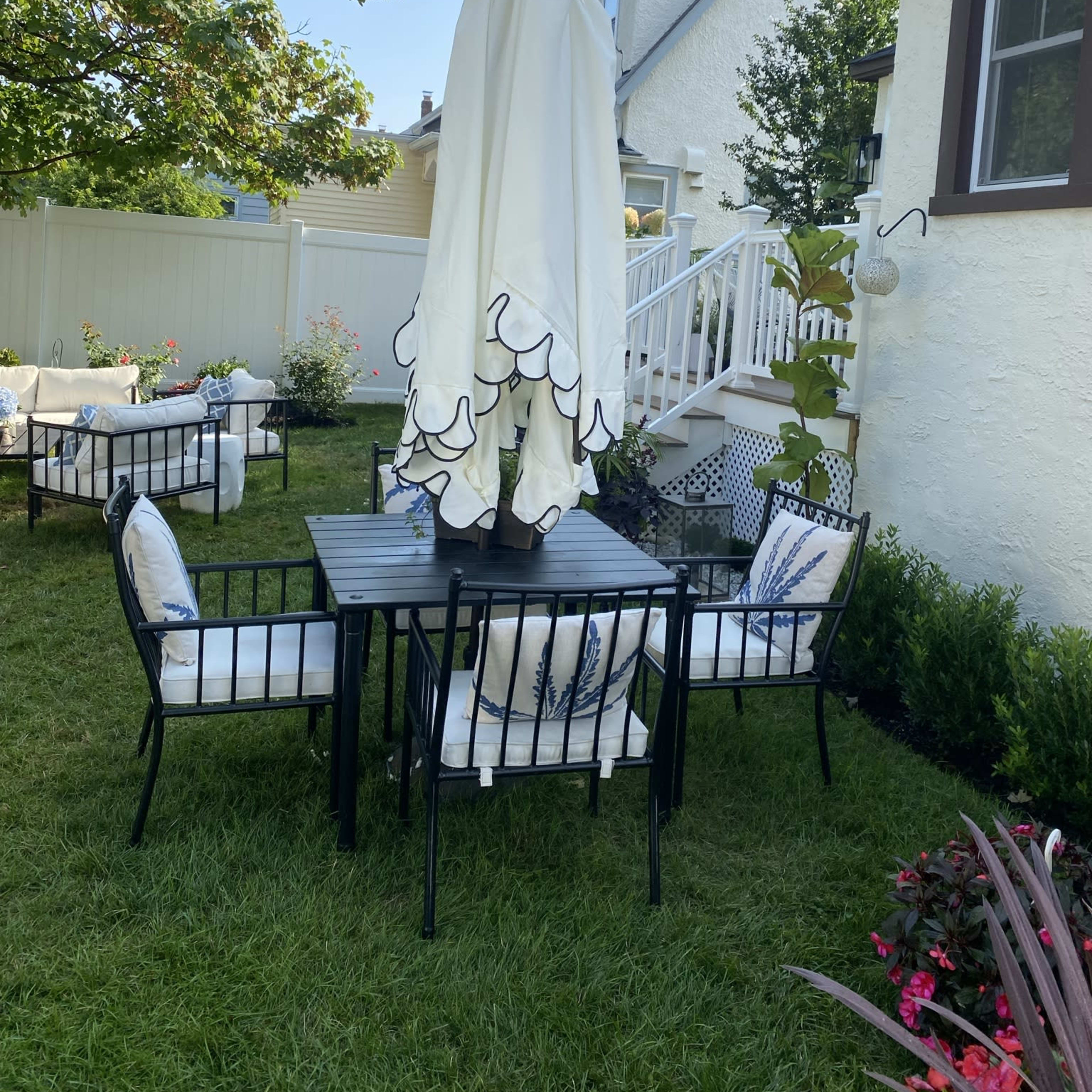 A black metal table with four chairs, covered by a large white umbrella, is set up on a grassy lawn beside a house.