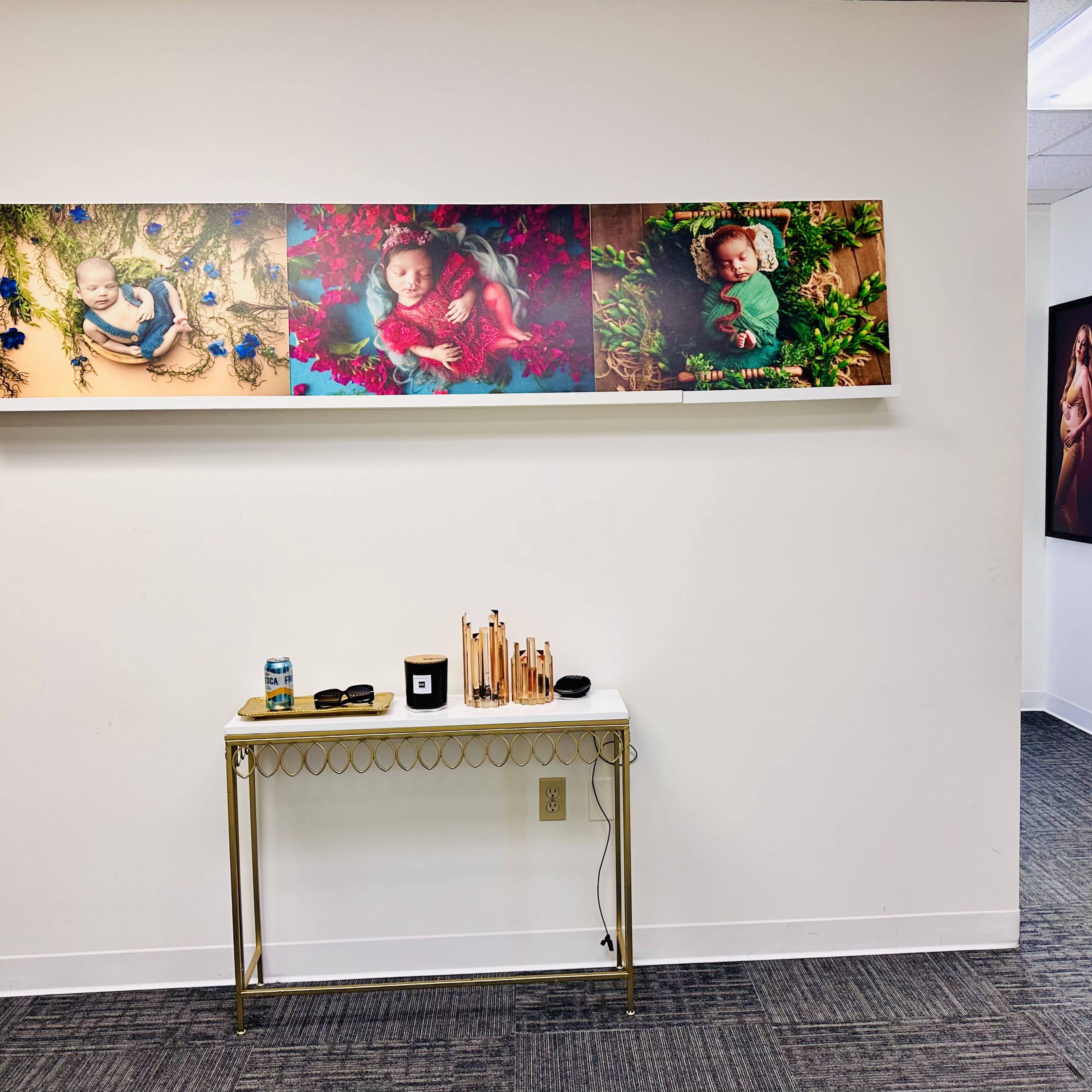 A wall with a decorative shelf displaying four photographs, while a table with various items, including a candle and sunglasses, is positioned below it in a room with gray carpet.