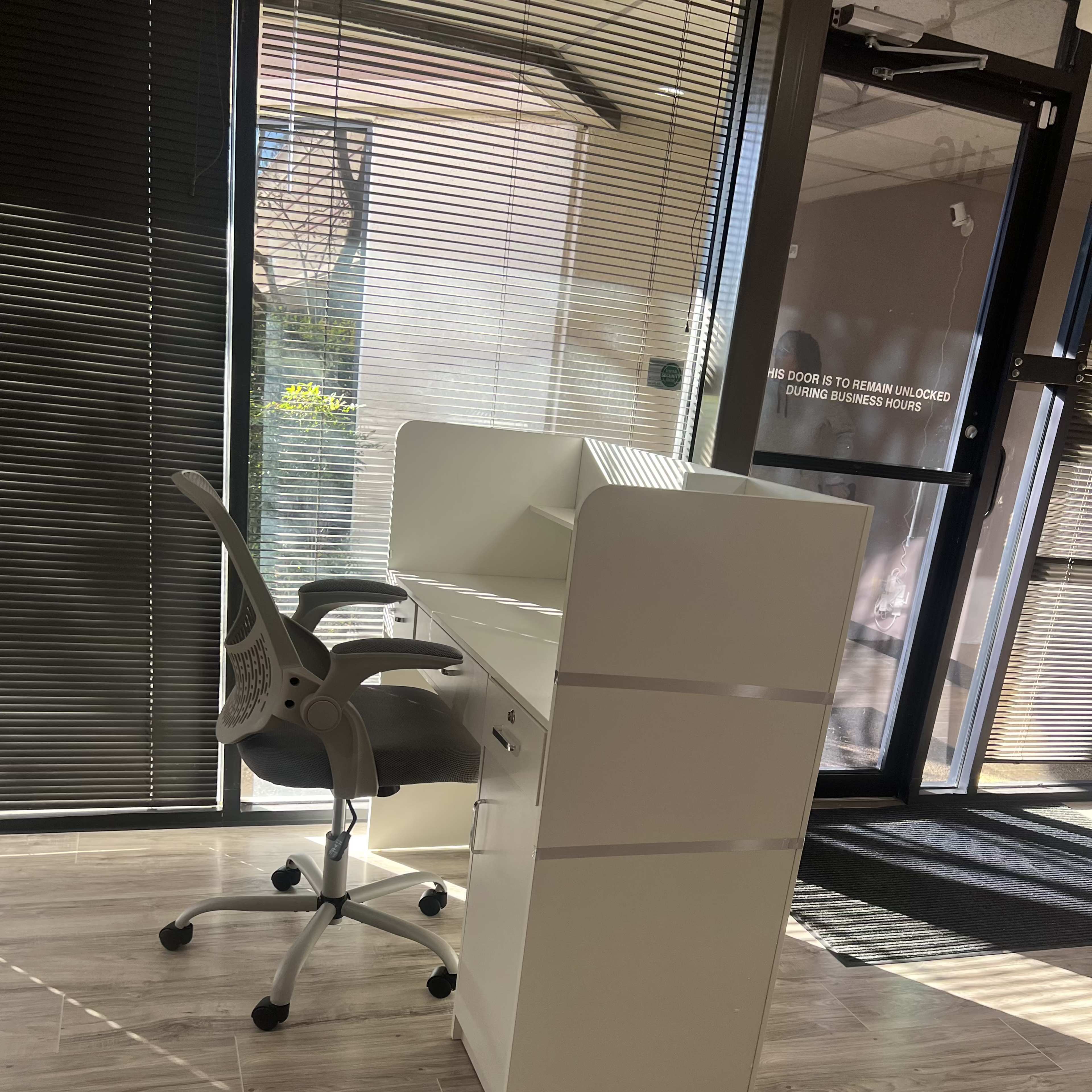 A modern white desk with a chair in a well-lit office space featuring large windows and wooden flooring.