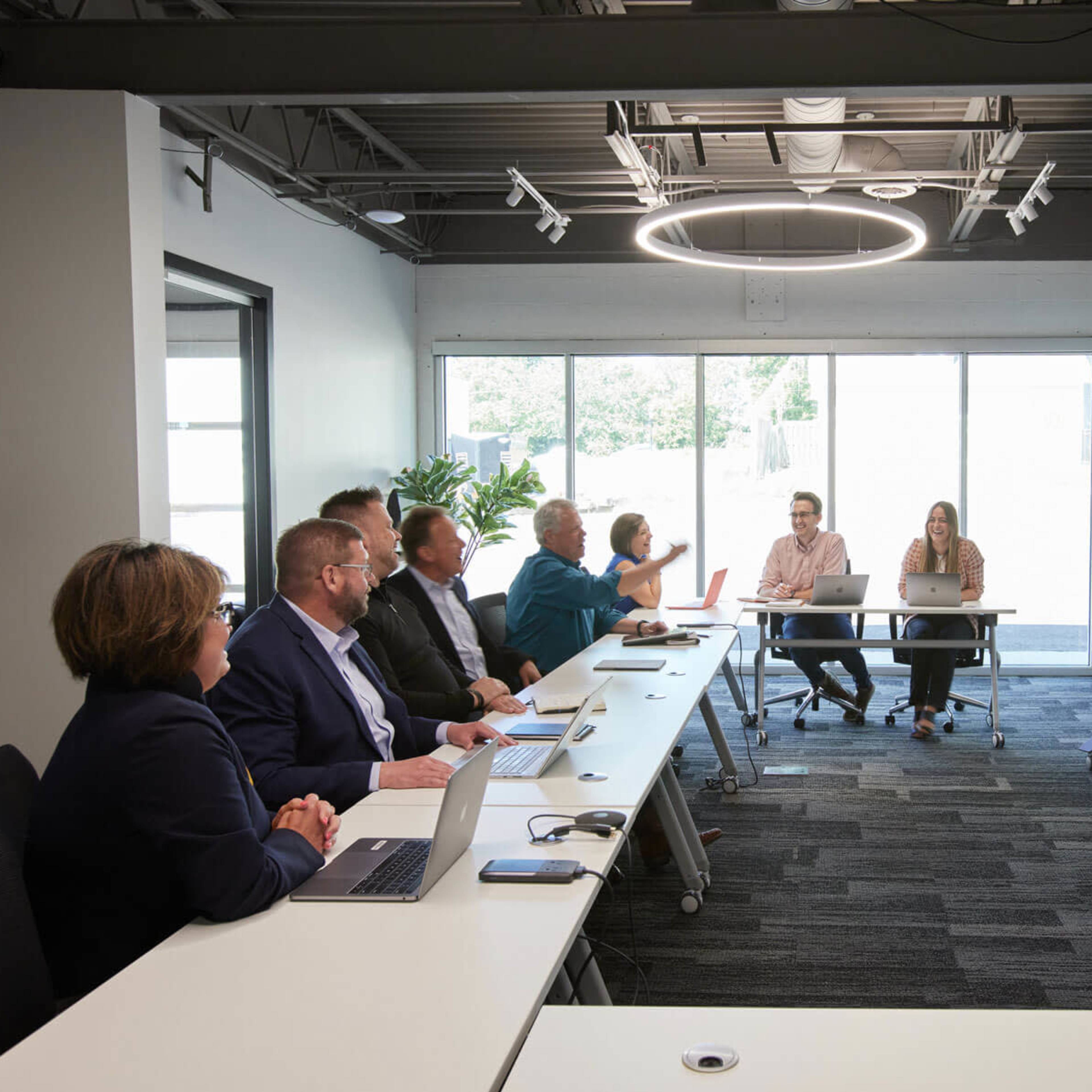 A group of eight professionals is seated at a long table in a modern conference room, while a woman stands at the front presenting to the team.
