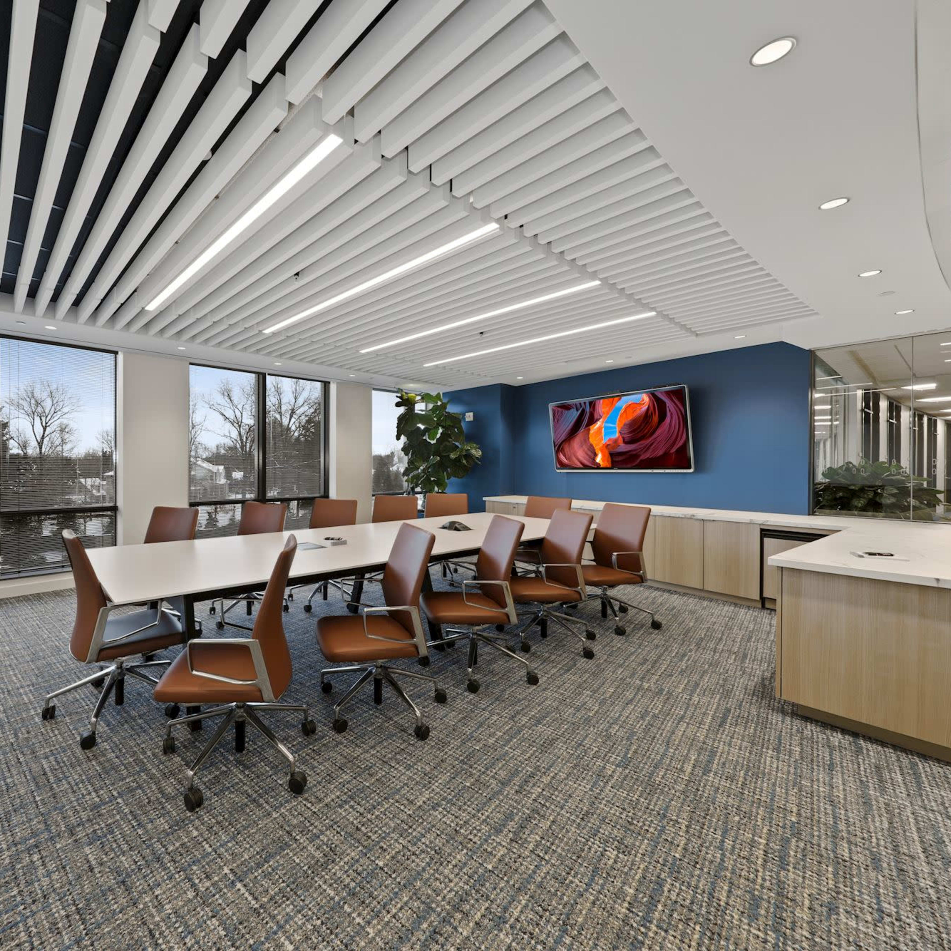 A modern conference room features a long table with rolling chairs, large windows, and a wall-mounted screen.
