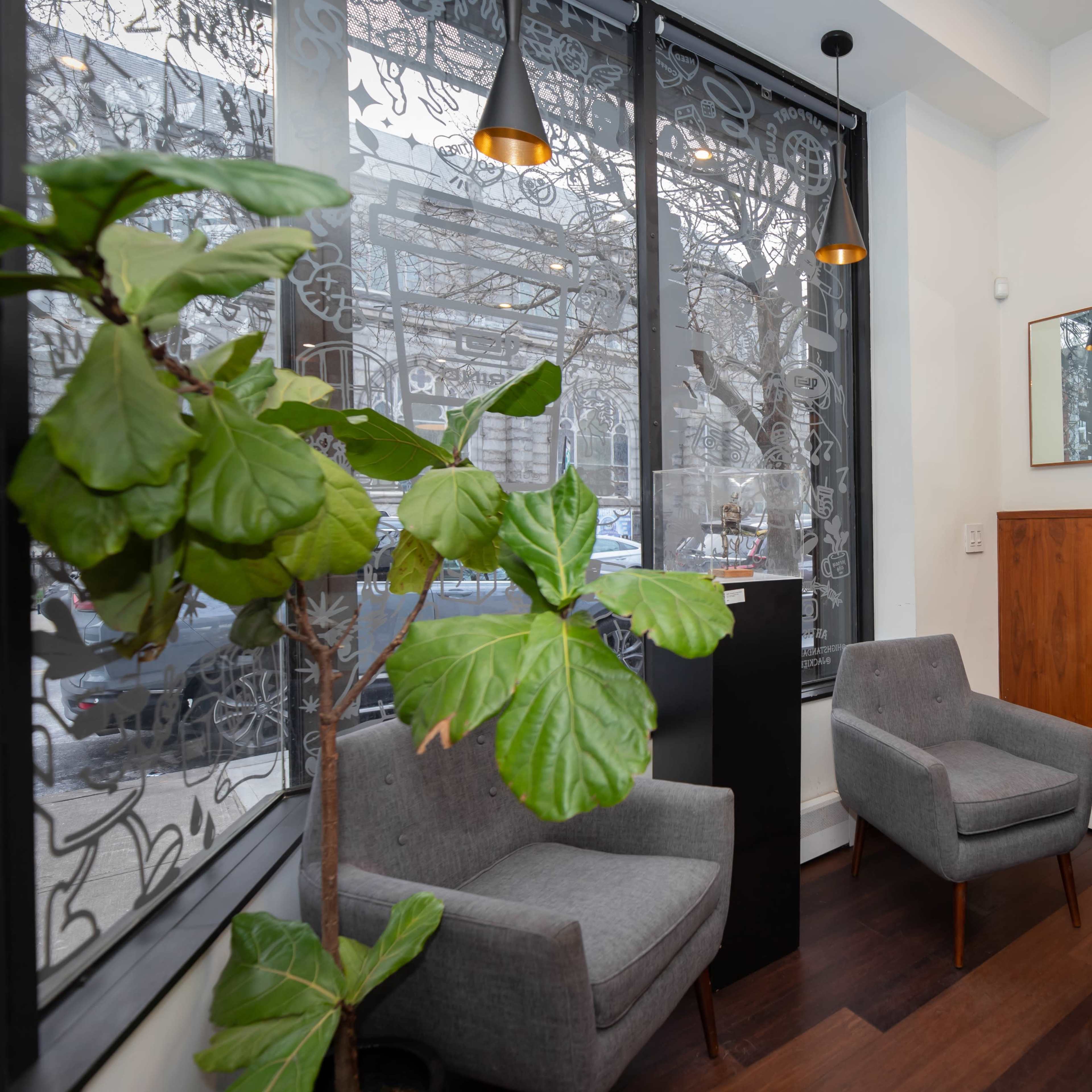 A cozy café interior features two gray chairs, a wooden table, and large windows with textured curtains, alongside a potted plant and a mirrored wall decor.