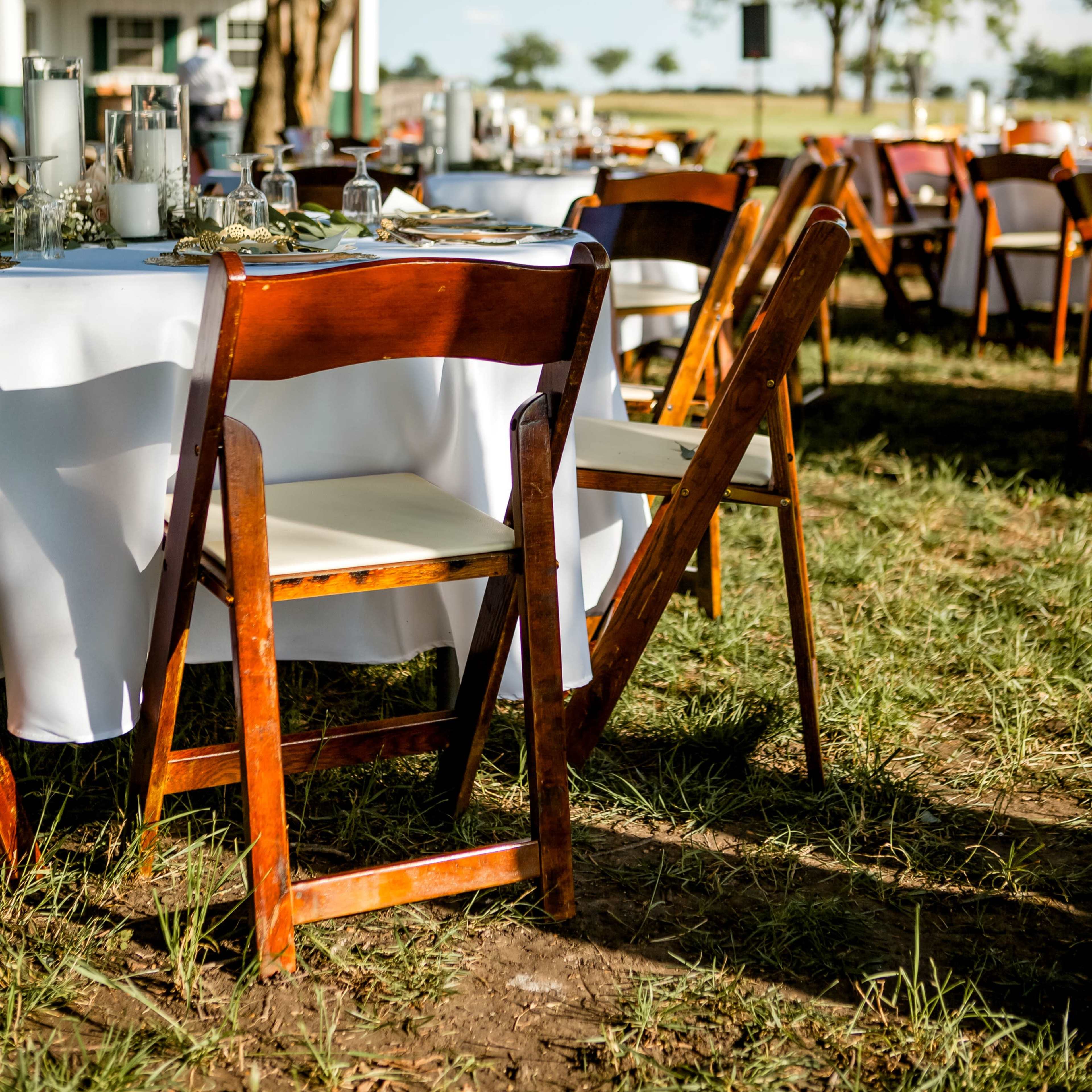 A rustic outdoor dining setup with wooden tables and chairs arranged on grass, beneath a clear sky.
