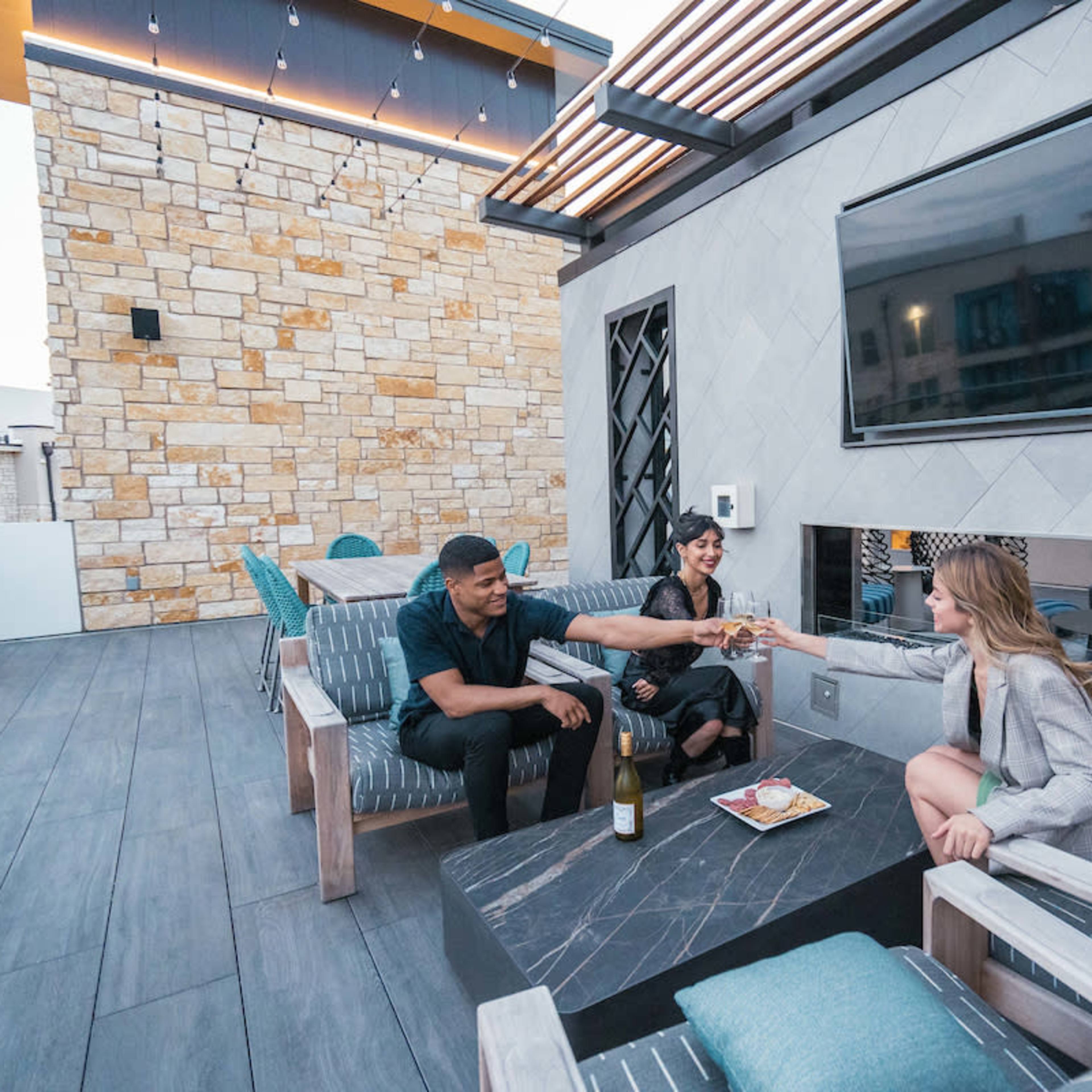 Three people are seated on a deck area, toasting with glasses near a stone wall and a large television screen.