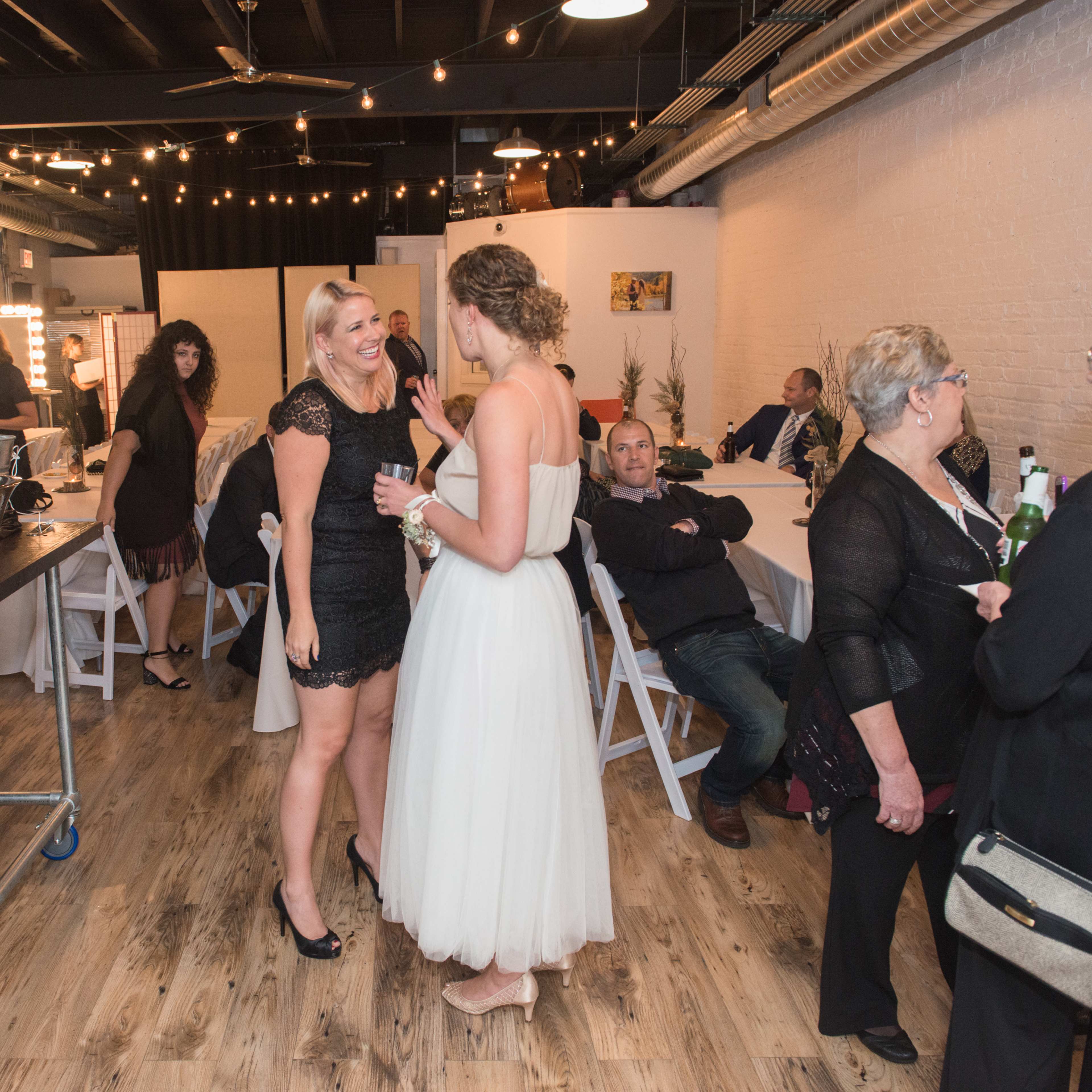 A group of people socialize in a decorated event space with tables and chairs, while two women converse in the foreground.