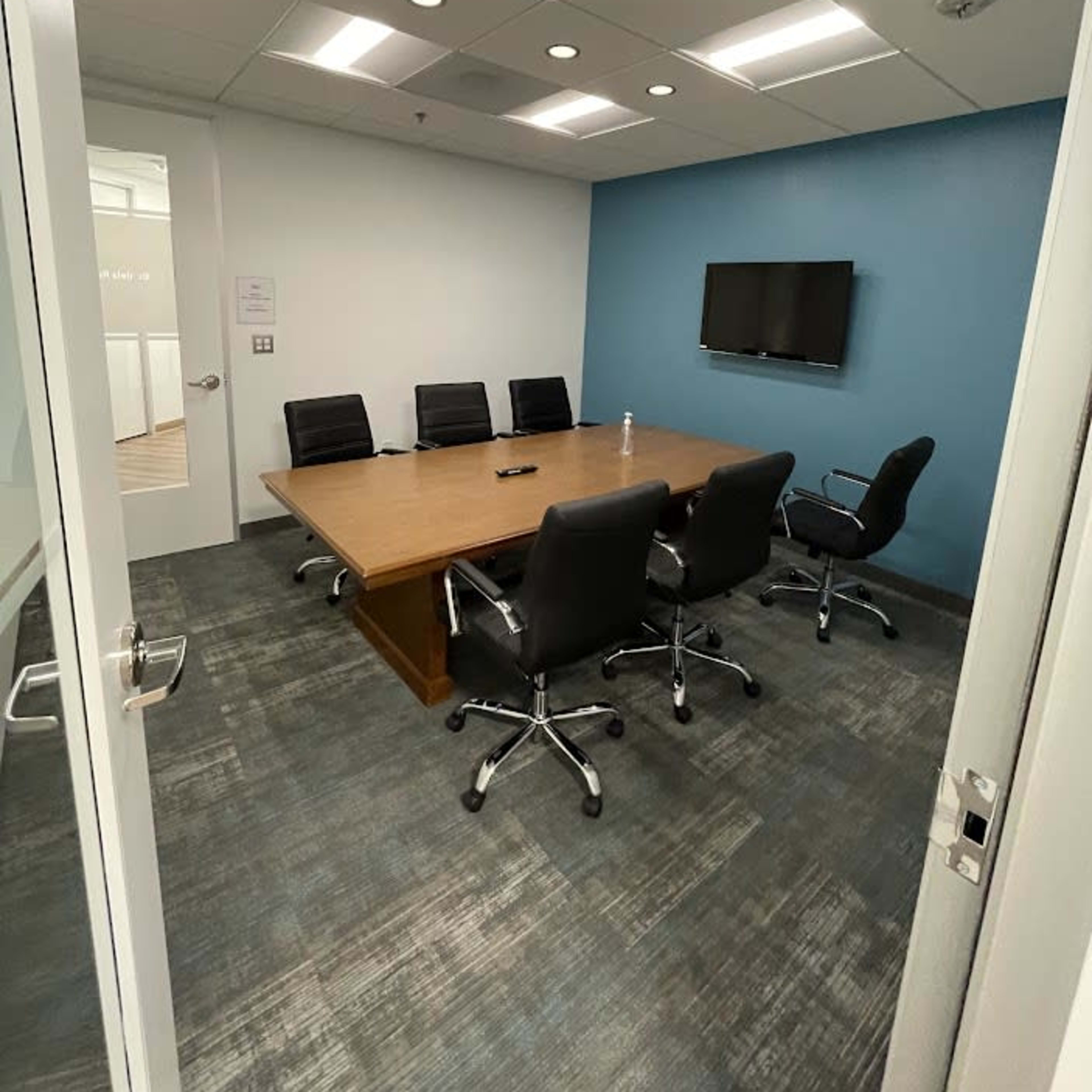 A meeting room features a large wooden table surrounded by black chairs, with a wall-mounted TV and blue accent wall.