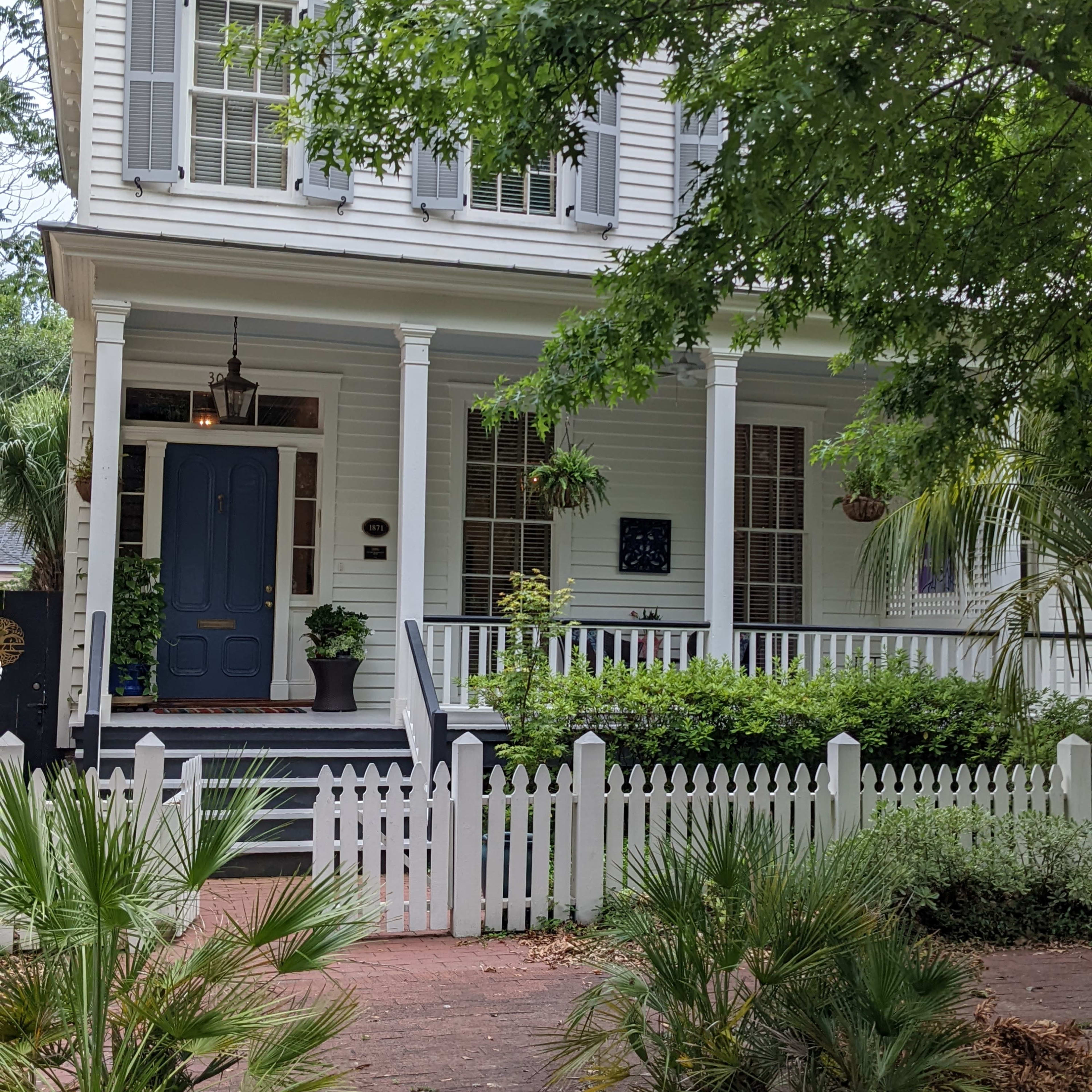 A white two-story house with a blue door, surrounded by greenery and a white picket fence.