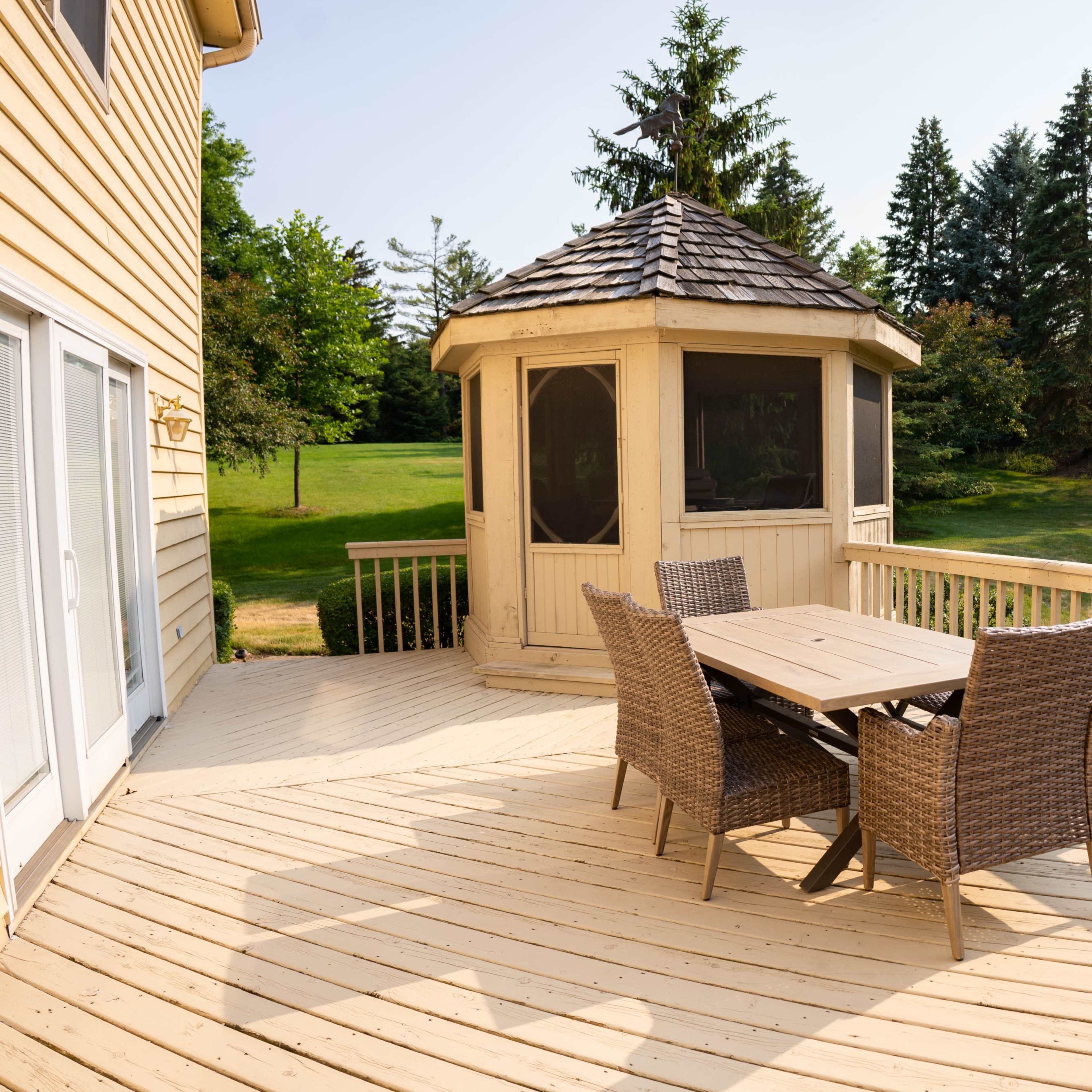 A wooden deck with a table and chairs overlooks a gazebo and a grassy area surrounded by trees.