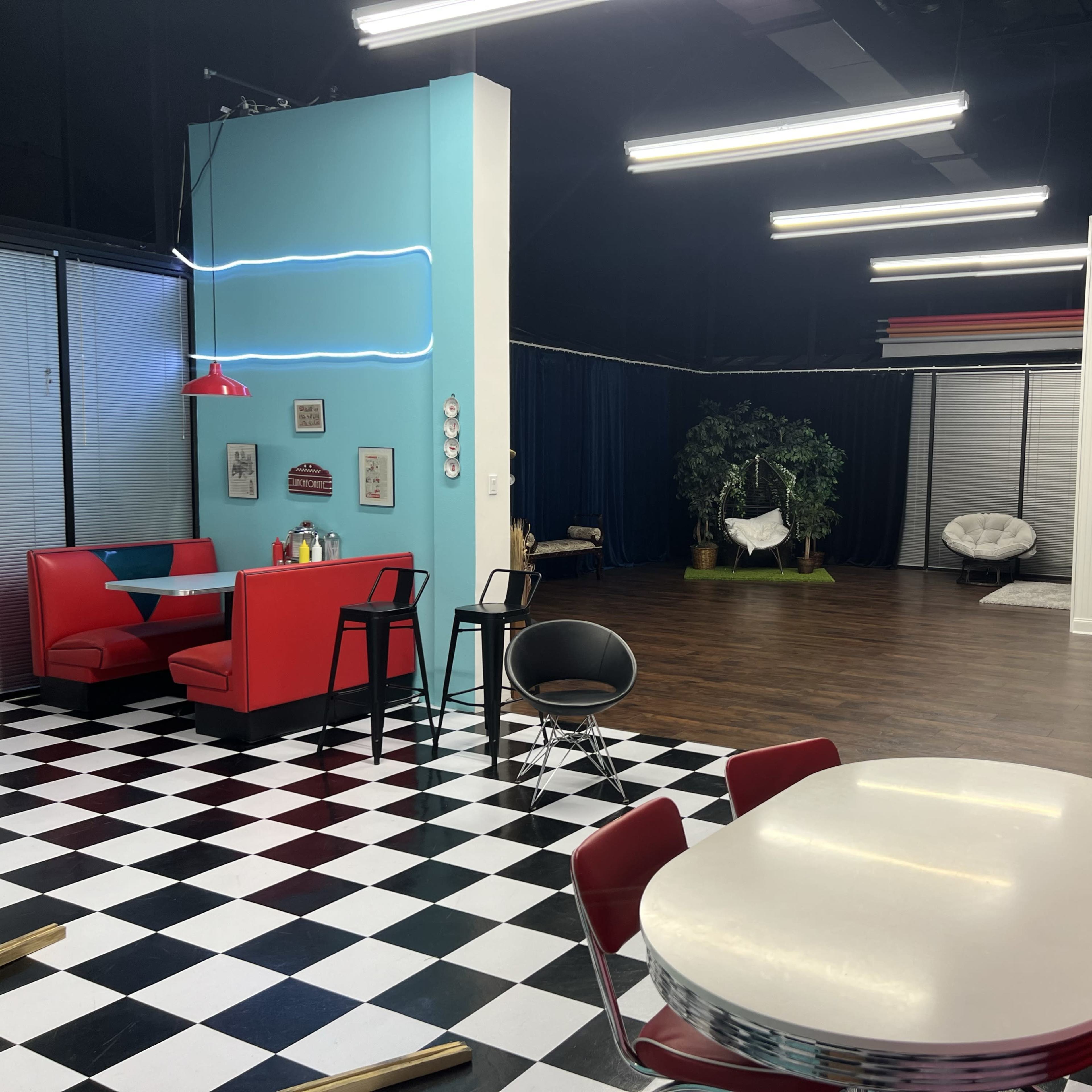 A colorful, modern interior featuring a diner-like seating area with red booths, a checkered floor, and a separate space in the background with plants and a hanging chair.