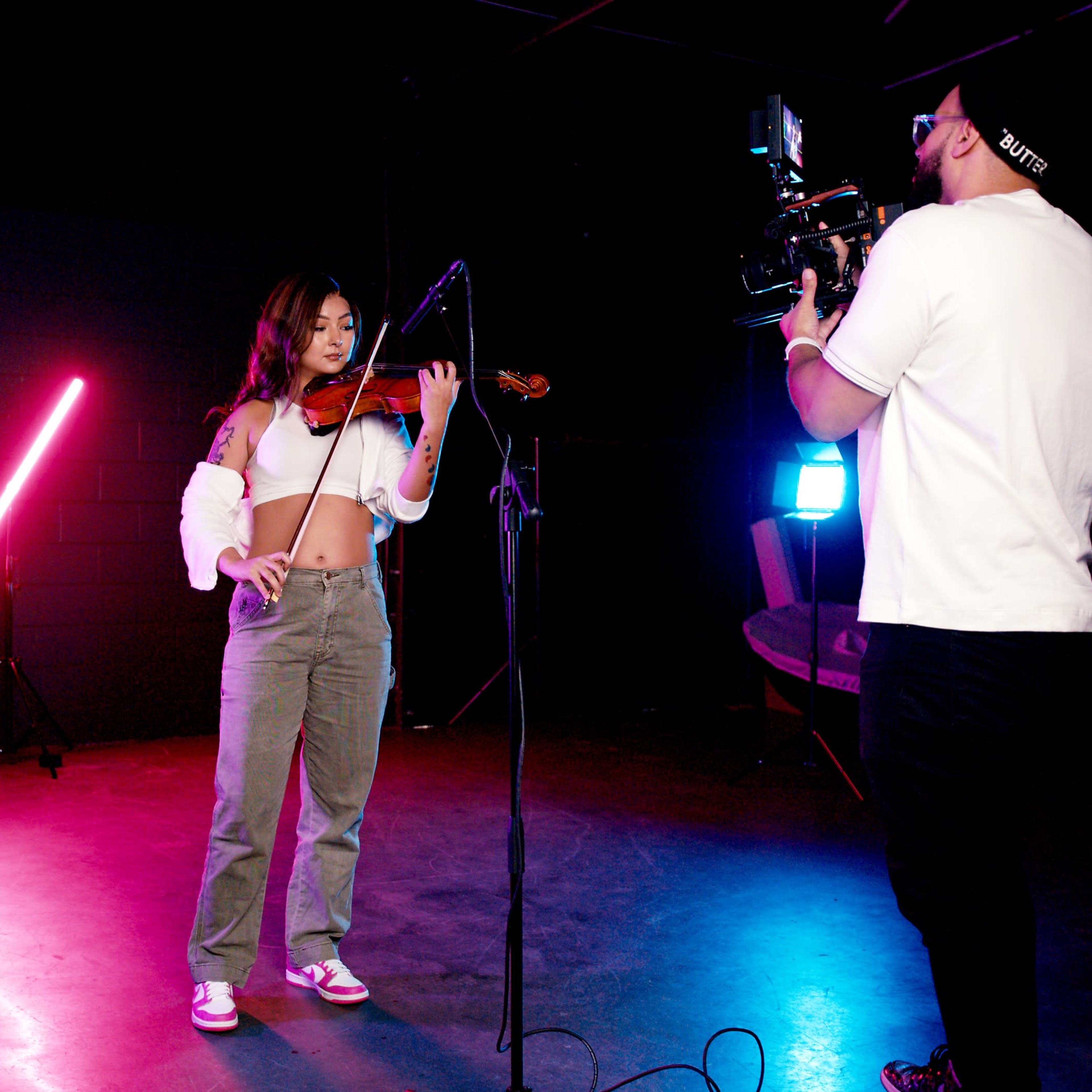 A young woman plays the violin while a man films her performance with a camera in a brightly lit studio with colored lights.