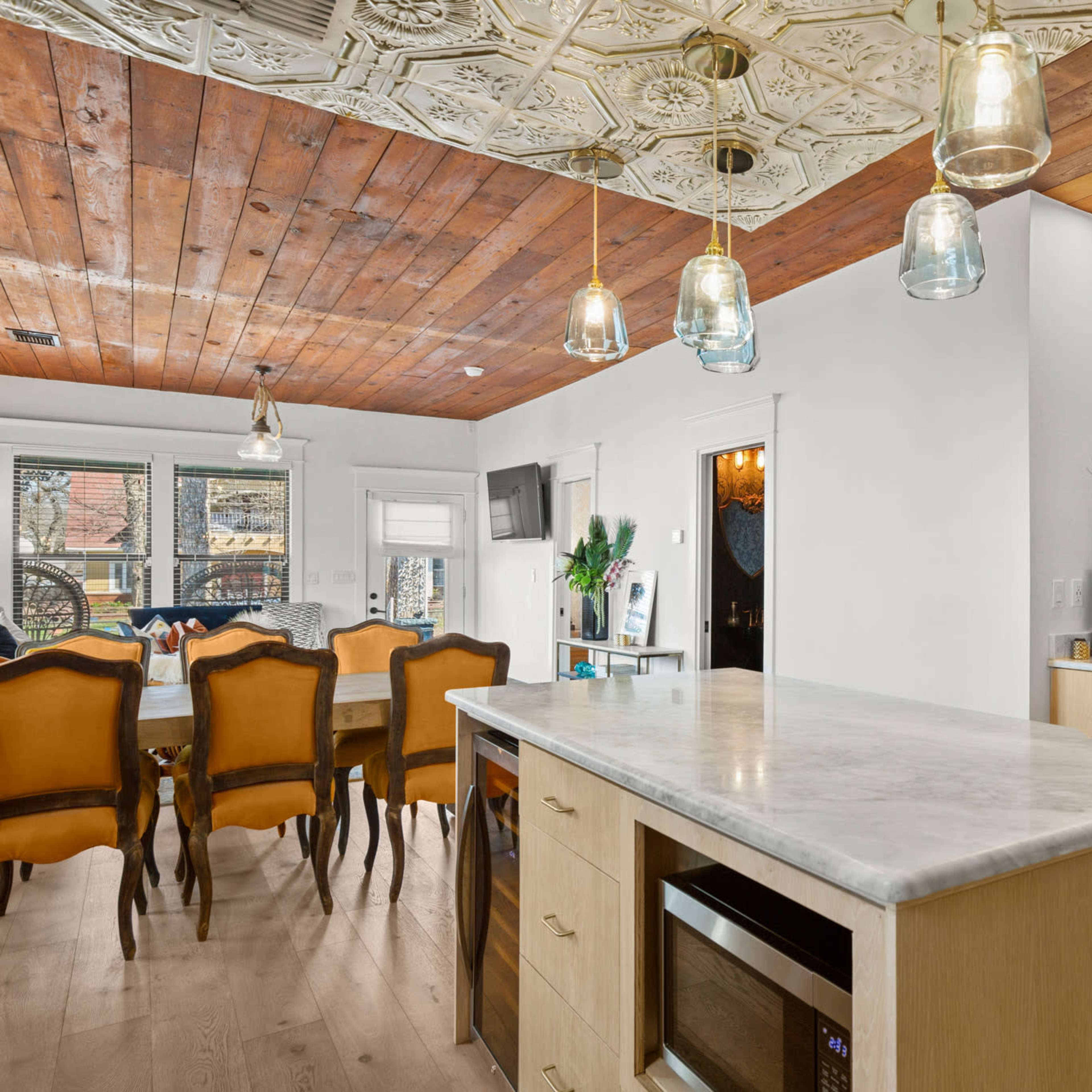 A modern kitchen with a large island featuring a marble countertop, pendant lights overhead, and a dining area in the background with a wooden ceiling and a table set for meals.