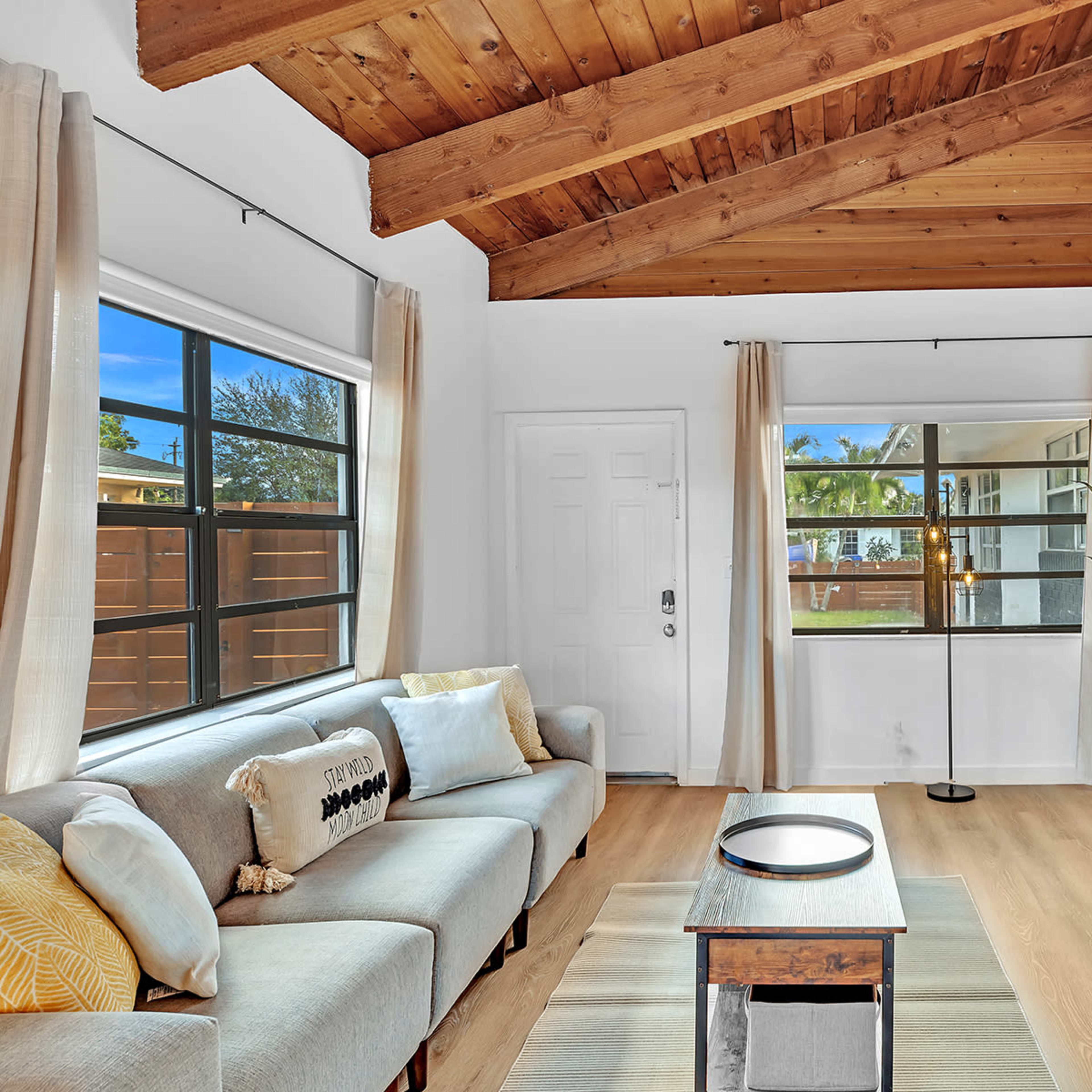 A living room features a sofa, a coffee table, a TV stand with a television, large windows, and a door leading outside, all under a wooden-beamed ceiling.