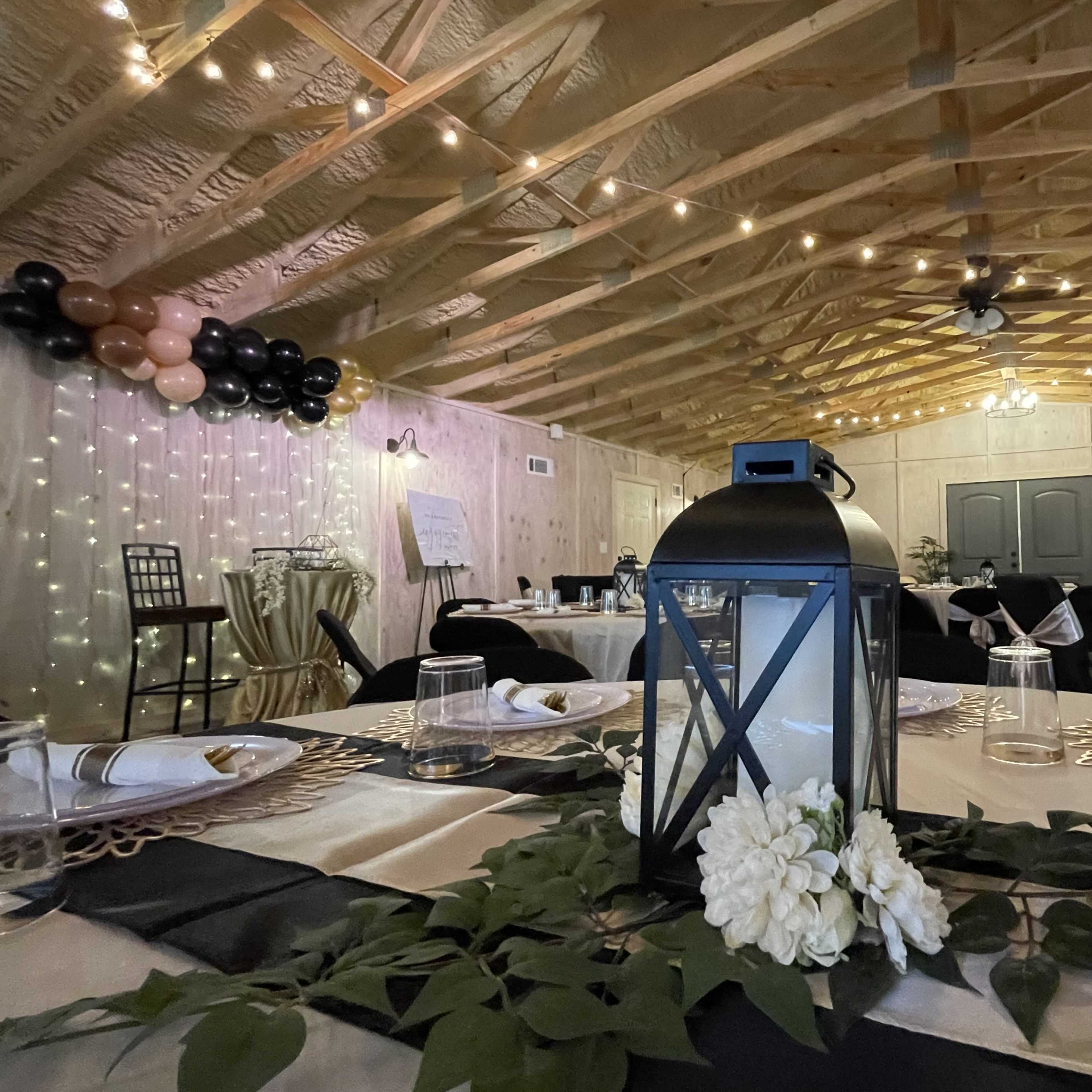 A decorated event space with tables set for a gathering, featuring a lantern centerpiece and string lights overhead.